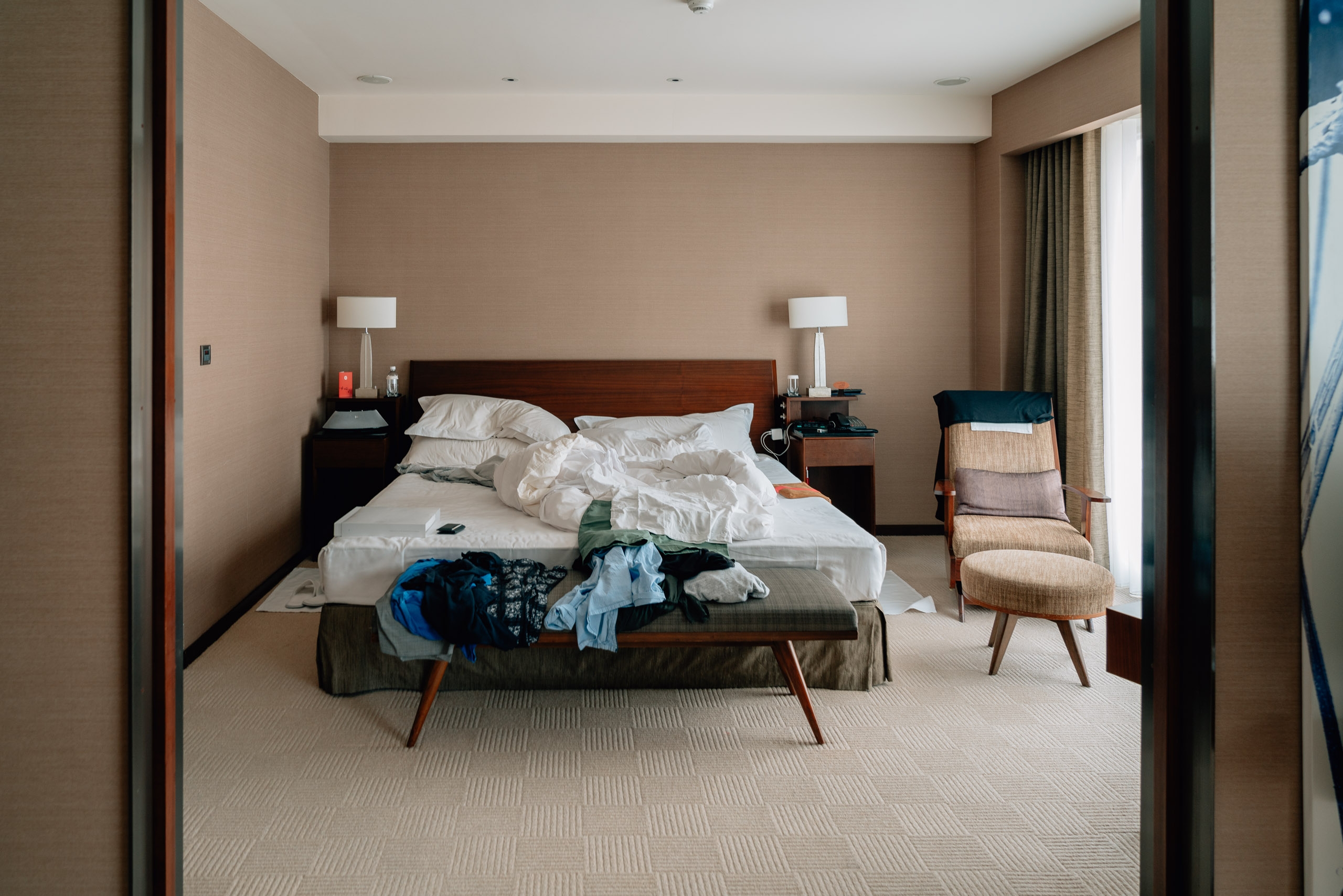 A modern hotel bedroom with taupe walls features an unmade bed with white linens, dark wood nightstands with white lamps, and a bench at the foot piled with clothing, alongside a woven accent chair