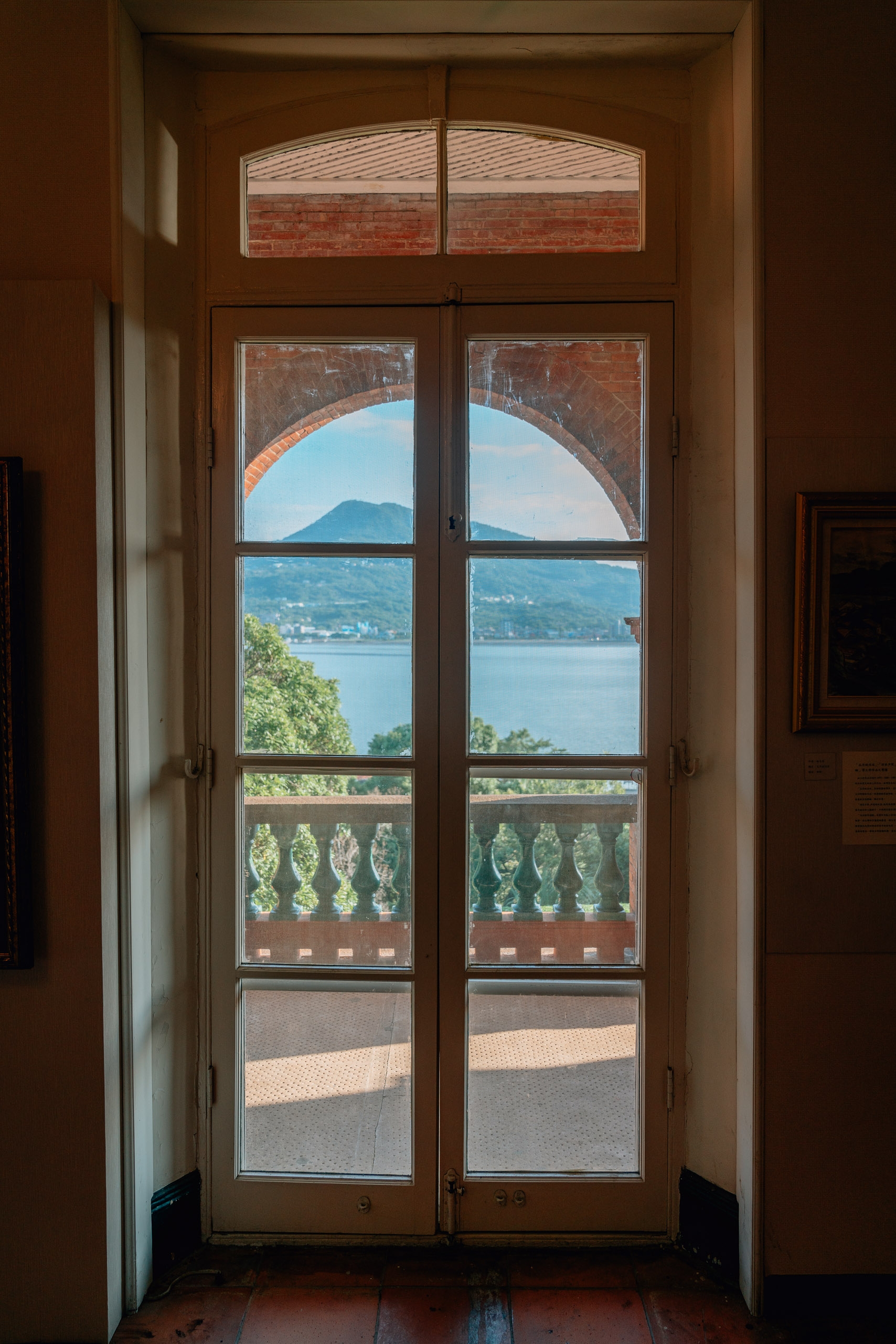 Double wooden doors frame a distant mountain and sparkling lake viewed through arched brick architecture and ornate balustrade railing in warm afternoon light