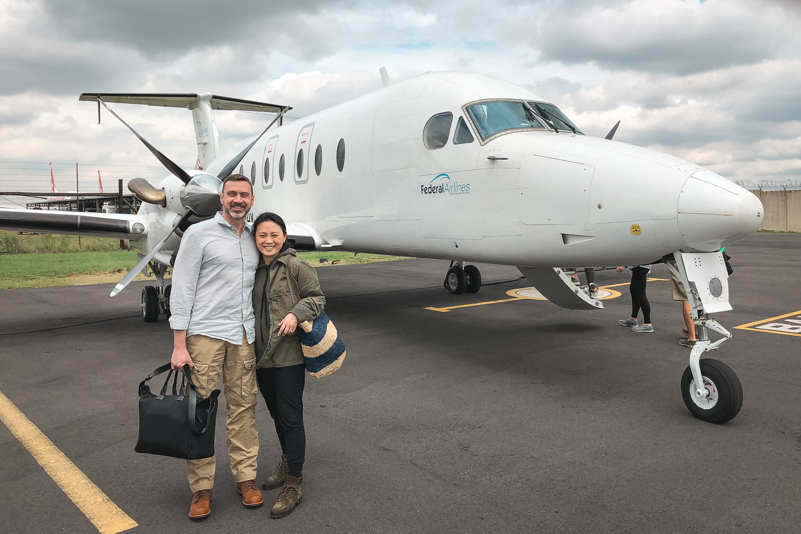 A man and woman stand together on a tarmac holding luggage beside a white twin-engine regional aircraft under overcast skies