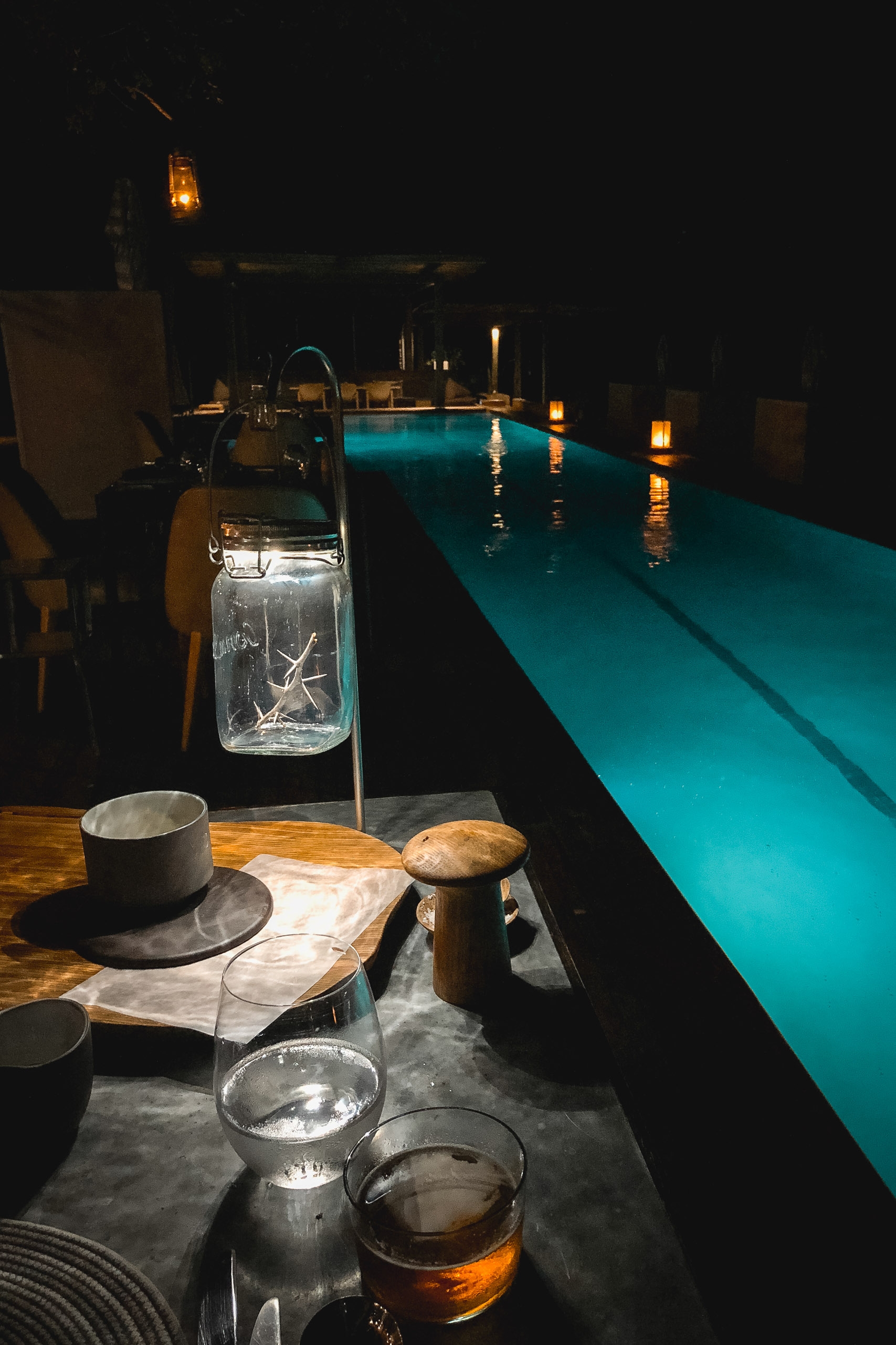 A nighttime poolside dining setup with turquoise-lit water, illuminated lanterns, and a table laden with bread, cups, and beverages overlooking a dark landscape