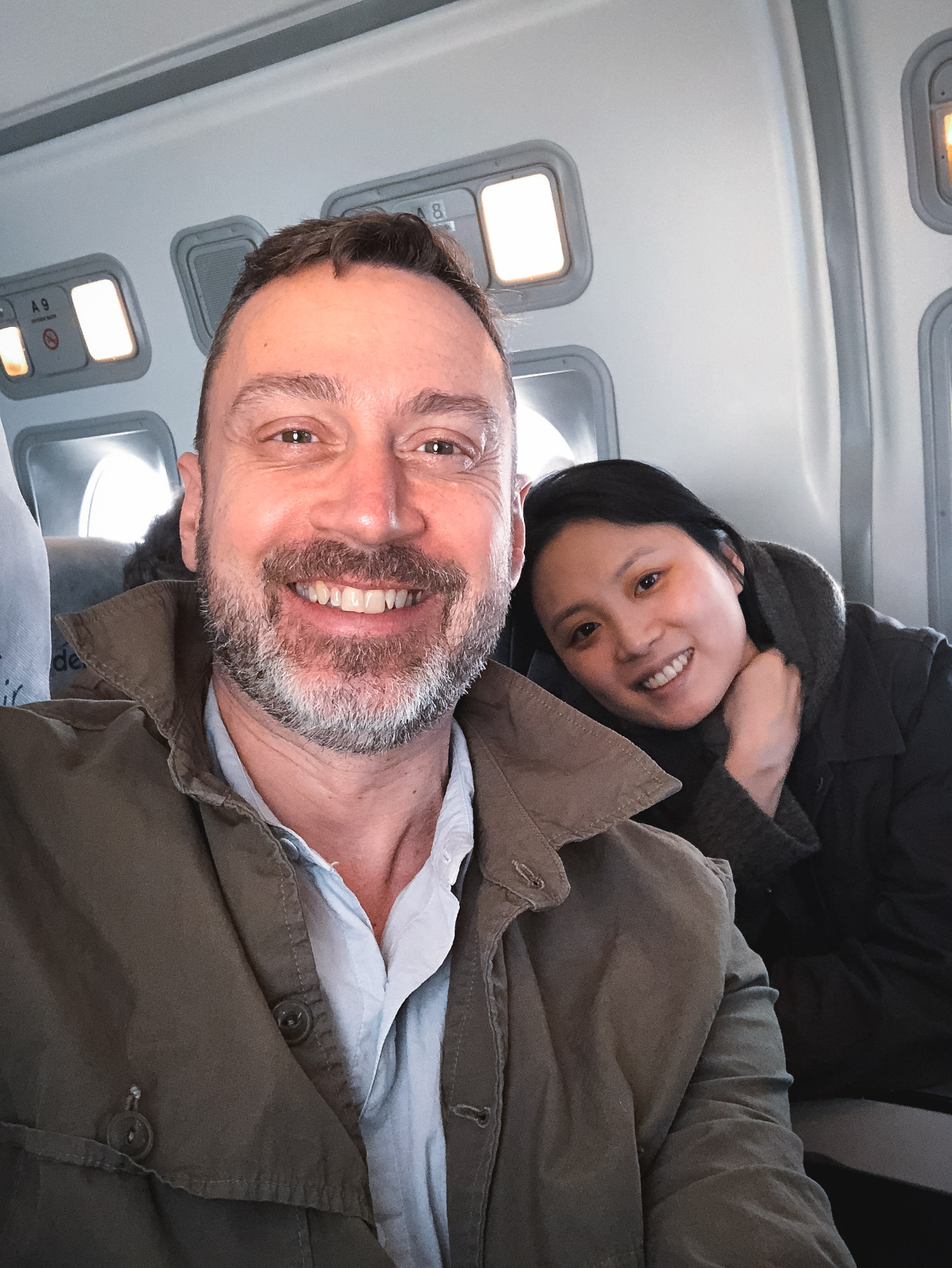 Two passengers smile at the camera inside a small aircraft cabin, with illuminated overhead panels and windows visible behind them