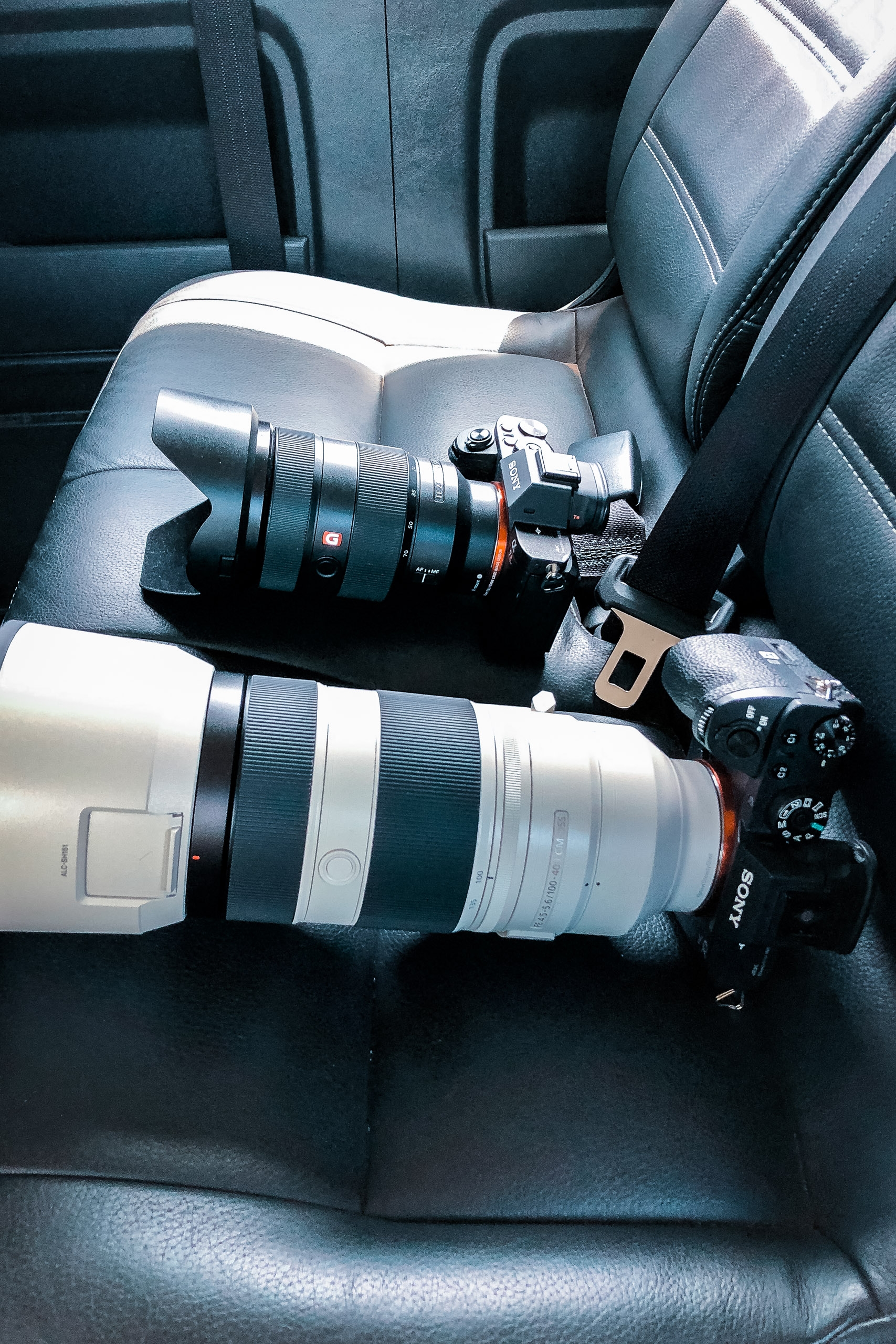 Professional camera equipment including two mirrorless cameras with telephoto lenses arranged on dark leather car seats in bright daylight