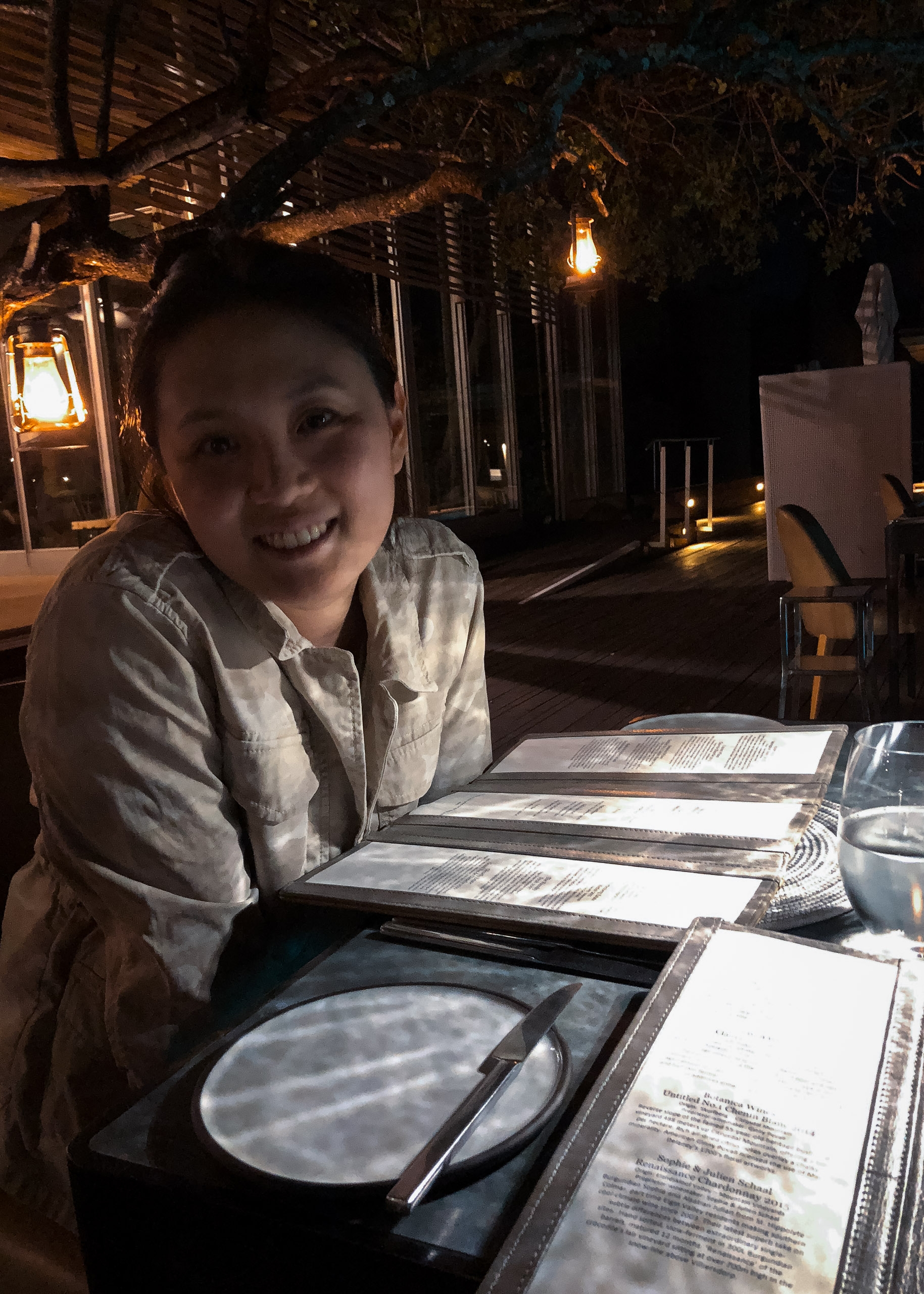 A woman smiles at a dining table set with place settings and menus under warm lantern lighting at an outdoor nighttime safari lodge with rustic wooden architecture overhead