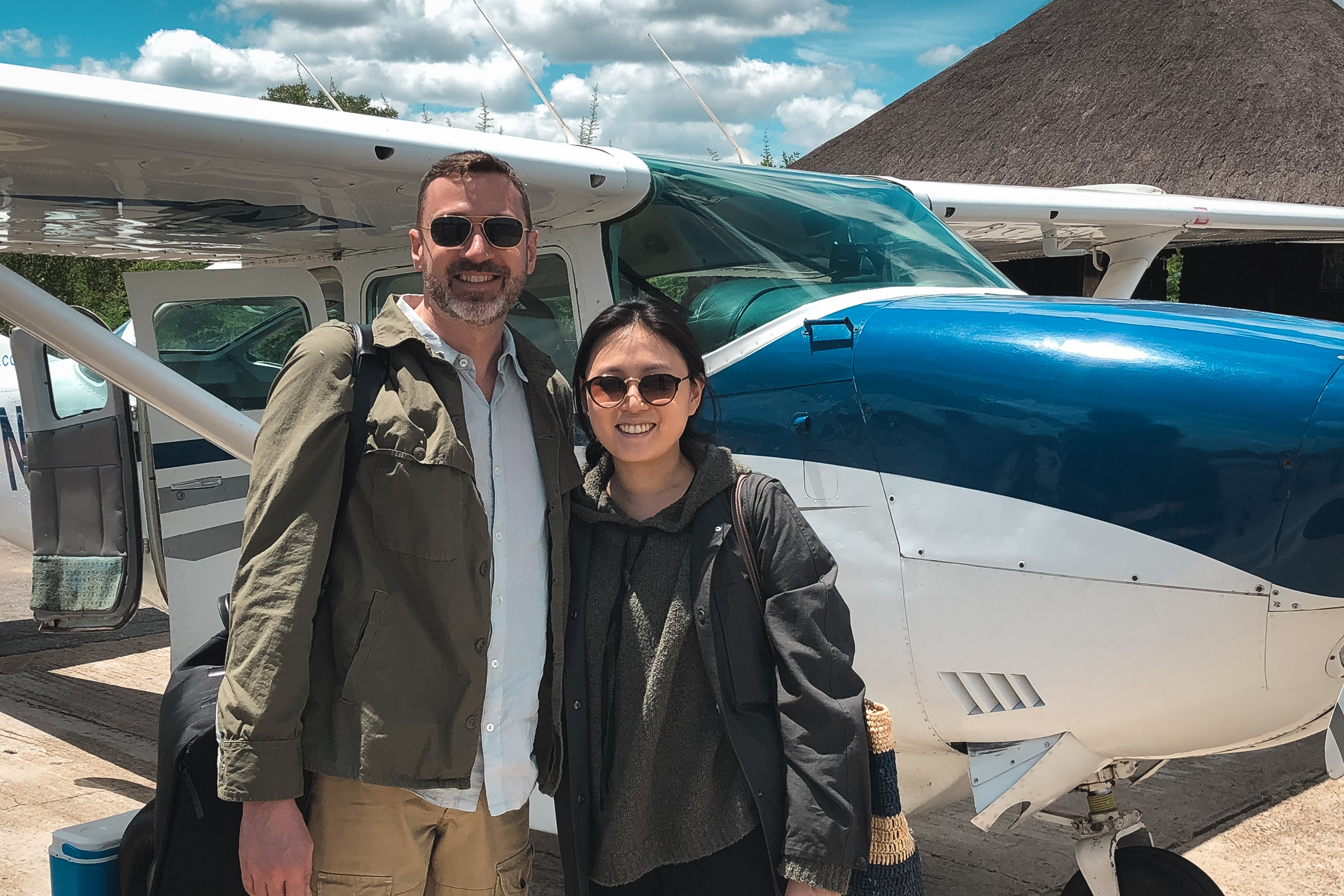 Two people stand smiling beside a blue and white aircraft on a sandy airstrip under partly cloudy skies, dressed in casual travel clothing