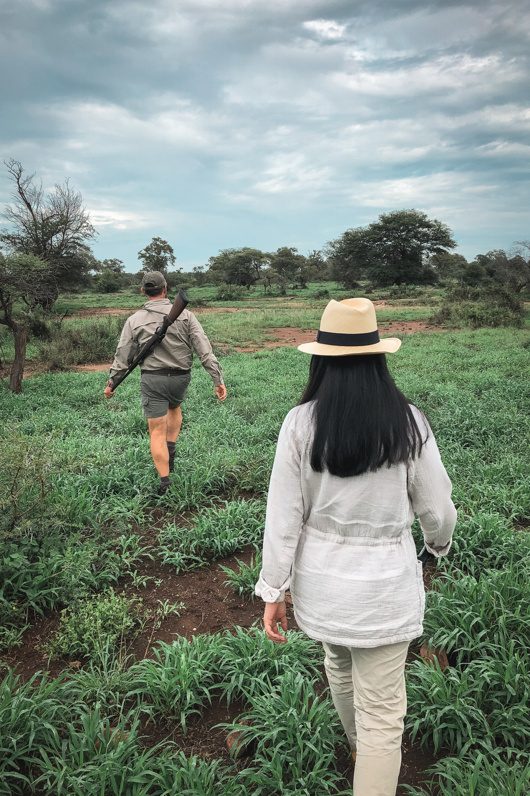 Two hikers walk away through a vast green savanna landscape with scattered acacia trees under an overcast sky with dramatic cloud formations