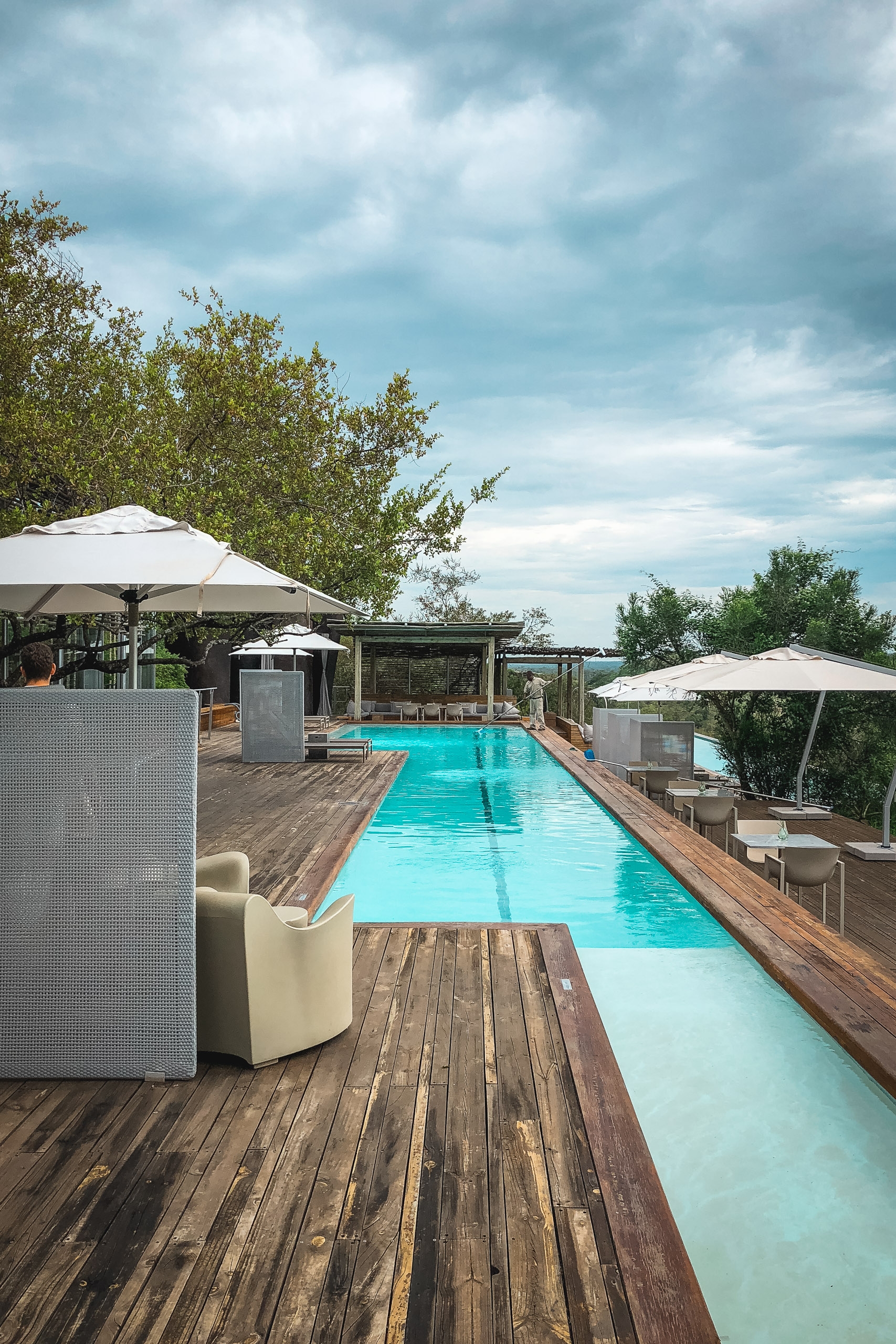 A modern rooftop pool with bright turquoise water stretches across a wooden deck, shaded by white umbrellas and surrounded by contemporary lounge furniture overlooking distant green landscape