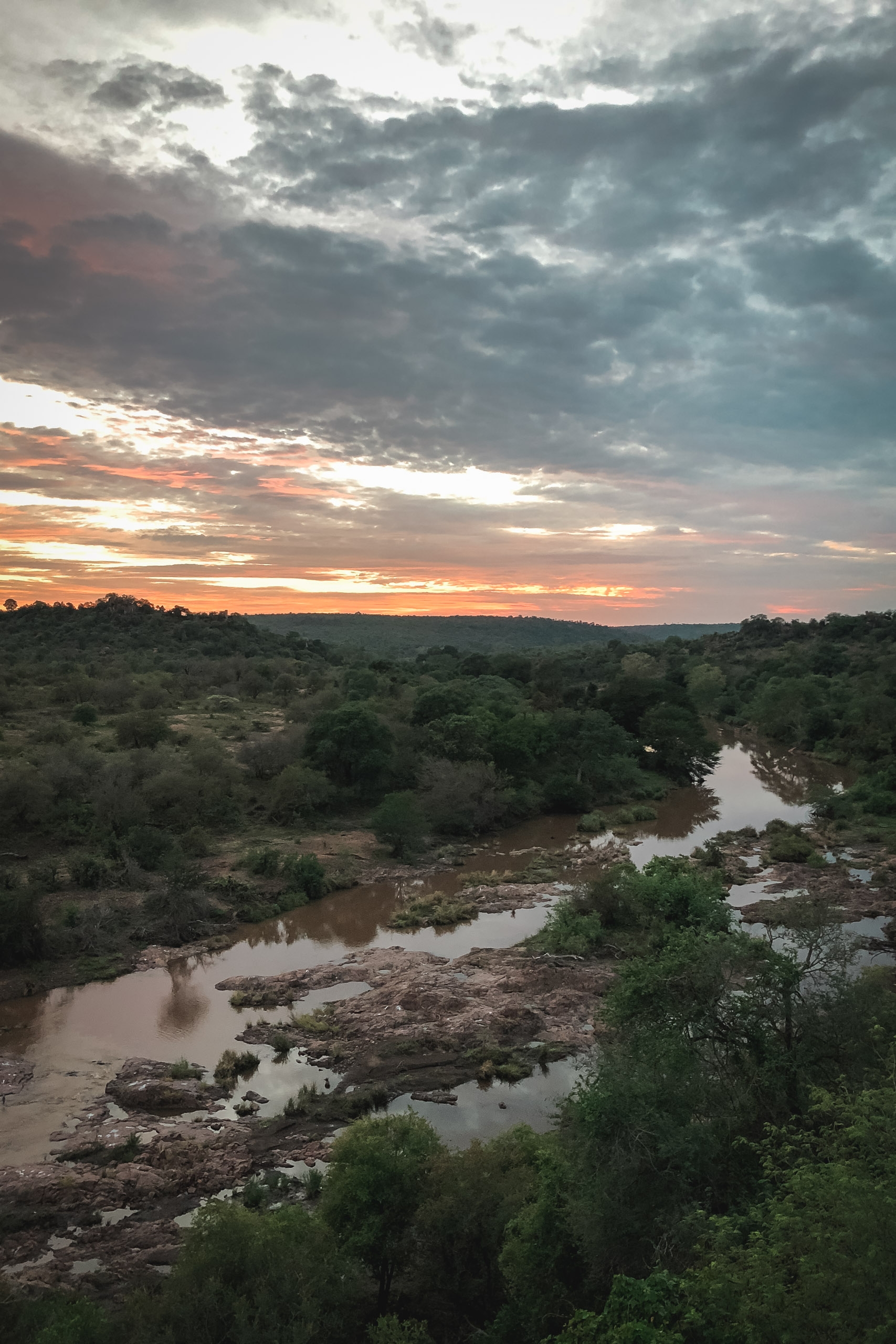 A winding river meanders through dense green bushveld landscape beneath a dramatic sunset sky with orange and gray storm clouds breaking across the horizon