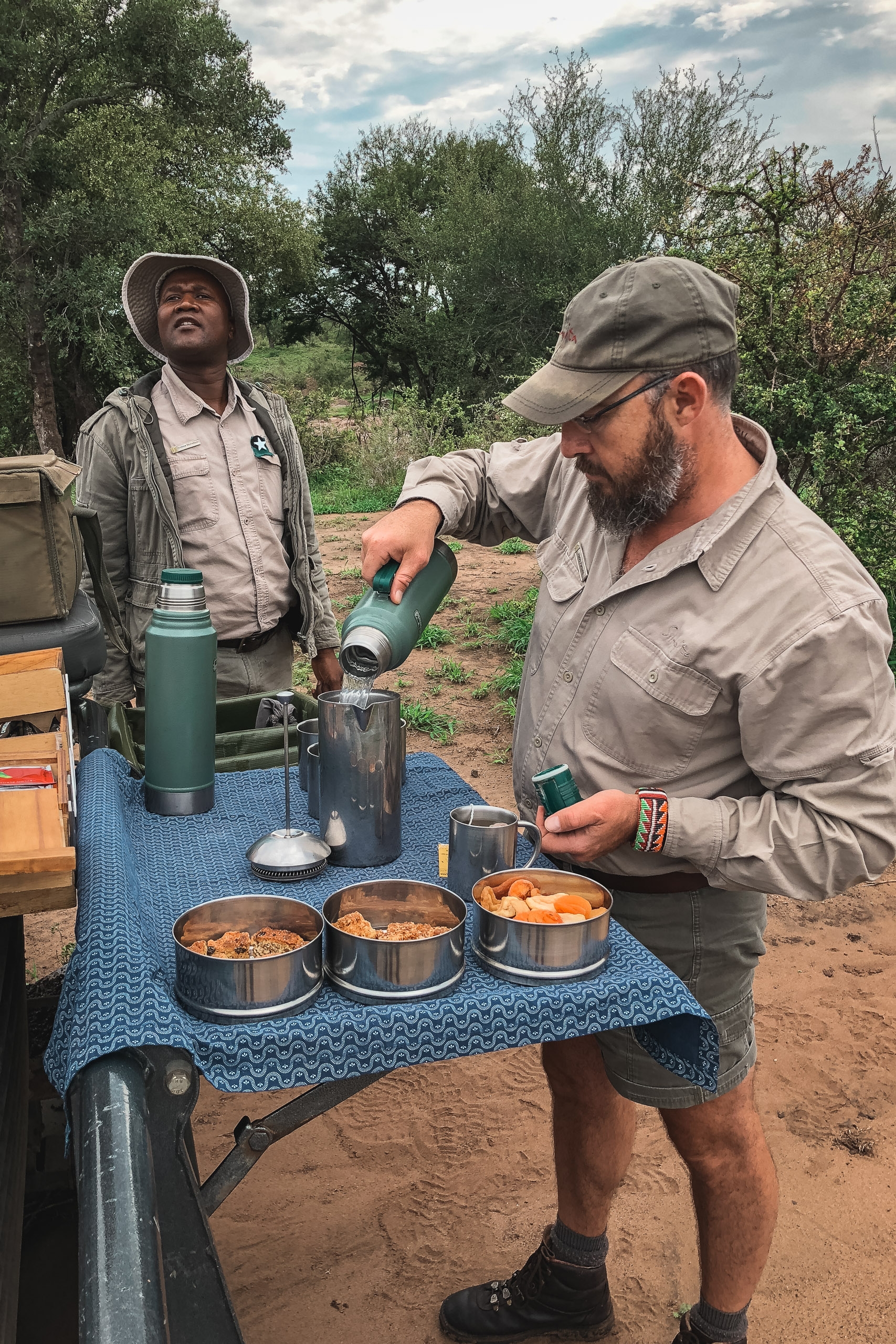 Two safari guides prepare a meal at a camp table with cooking pots, while trees and cloudy sky fill the background during daytime