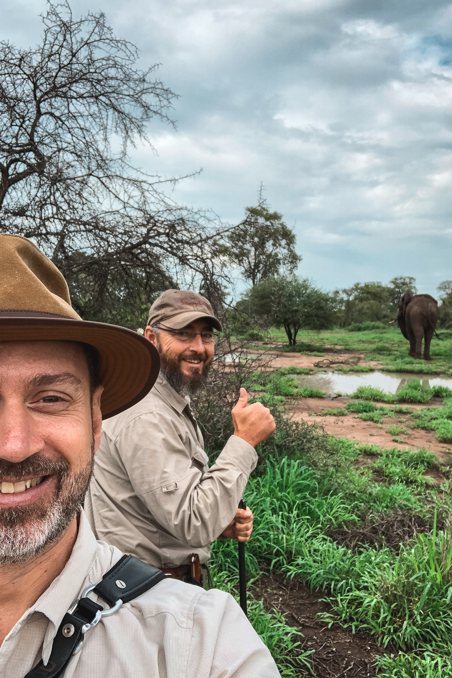 Two safari guides in khaki attire pose before an elephant in a lush African landscape with scattered trees, waterhole, and cloudy sky overhead