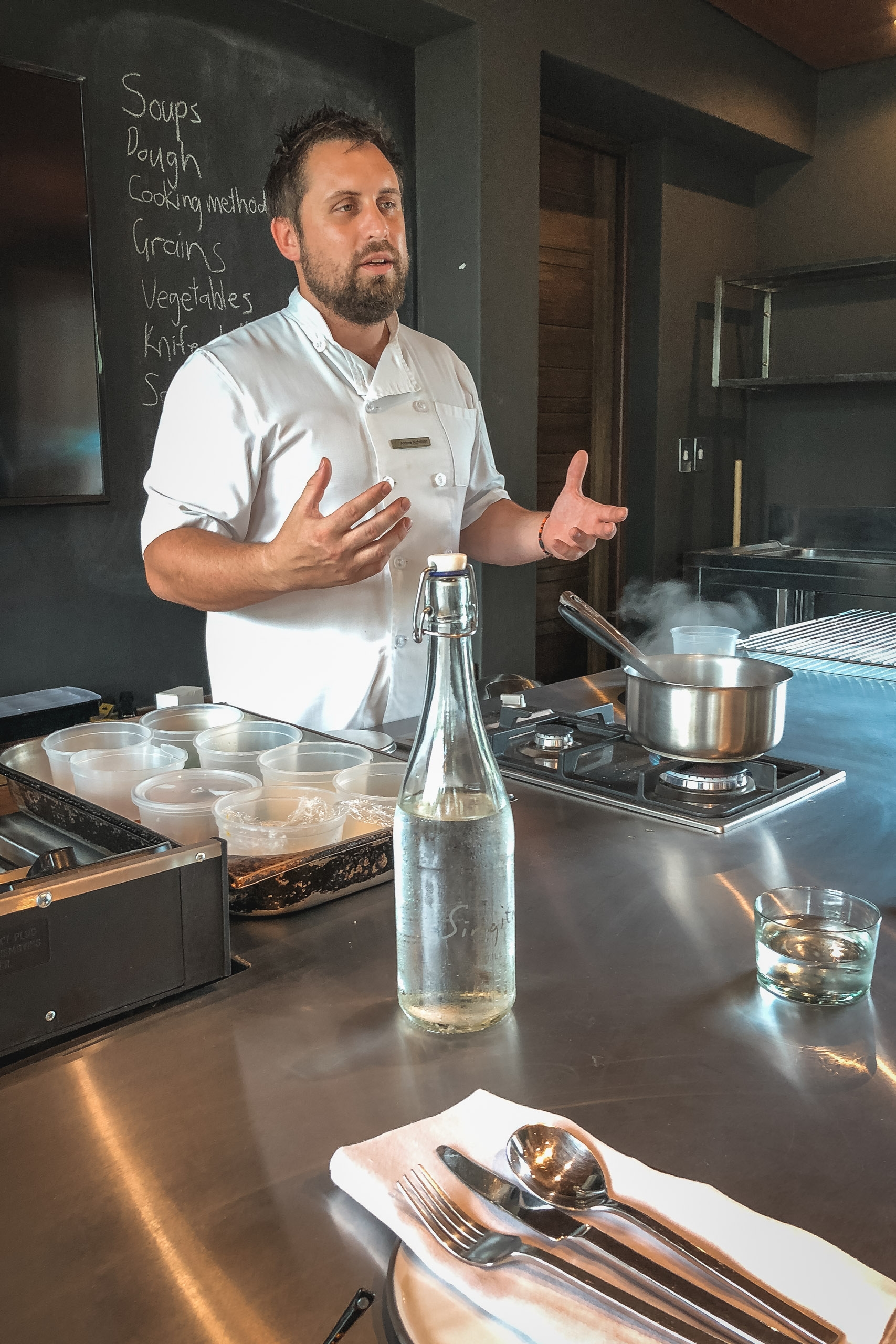 A bearded chef in a white chef's coat gestures expressively while conducting a cooking demonstration in a modern kitchen, with ingredient bowls and a pot on the counter behind him.