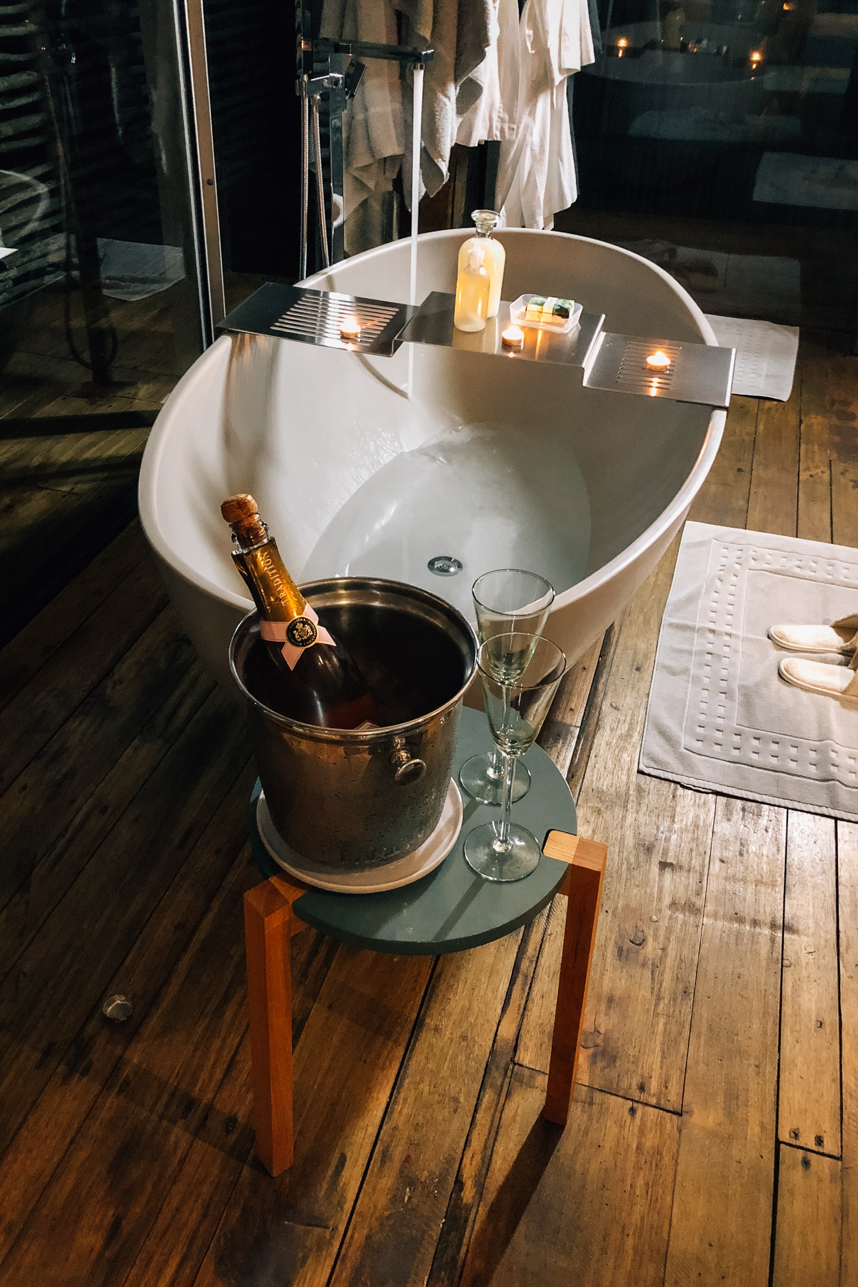 A luxurious spa bathtub with champagne bucket and glass on a wooden floor beside candlelit amenities and hanging robes in soft afternoon light