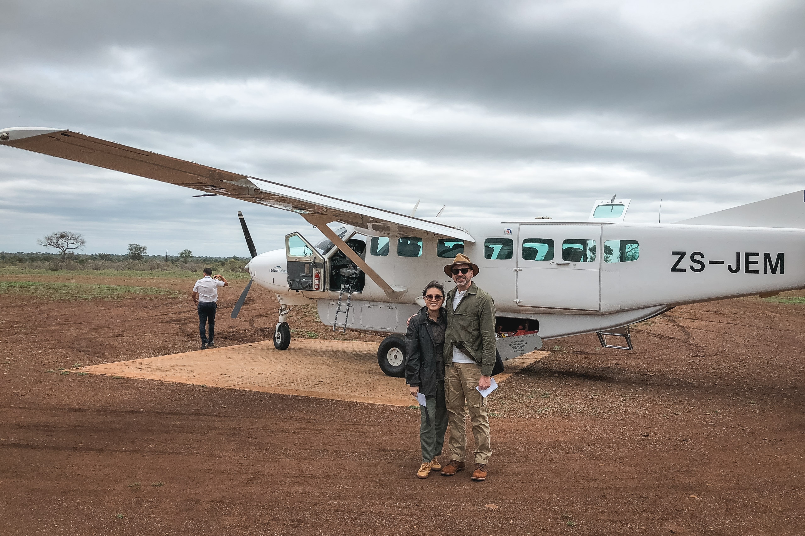 A small white charter plane with registration ZS-JEM sits on a red dirt airstrip in the African bush, with two people posing in front and overcast skies overhead