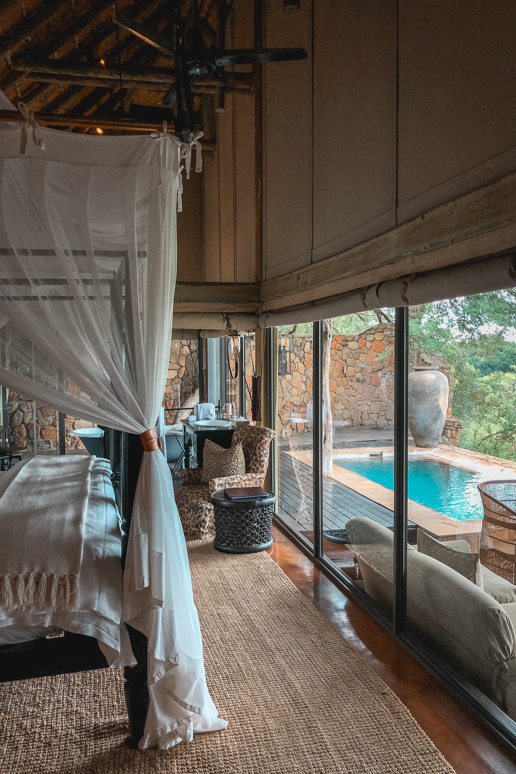 A luxury safari bedroom with sheer white curtains frames a bed overlooking a turquoise pool and stone walls, natural light streaming through expansive glass doors