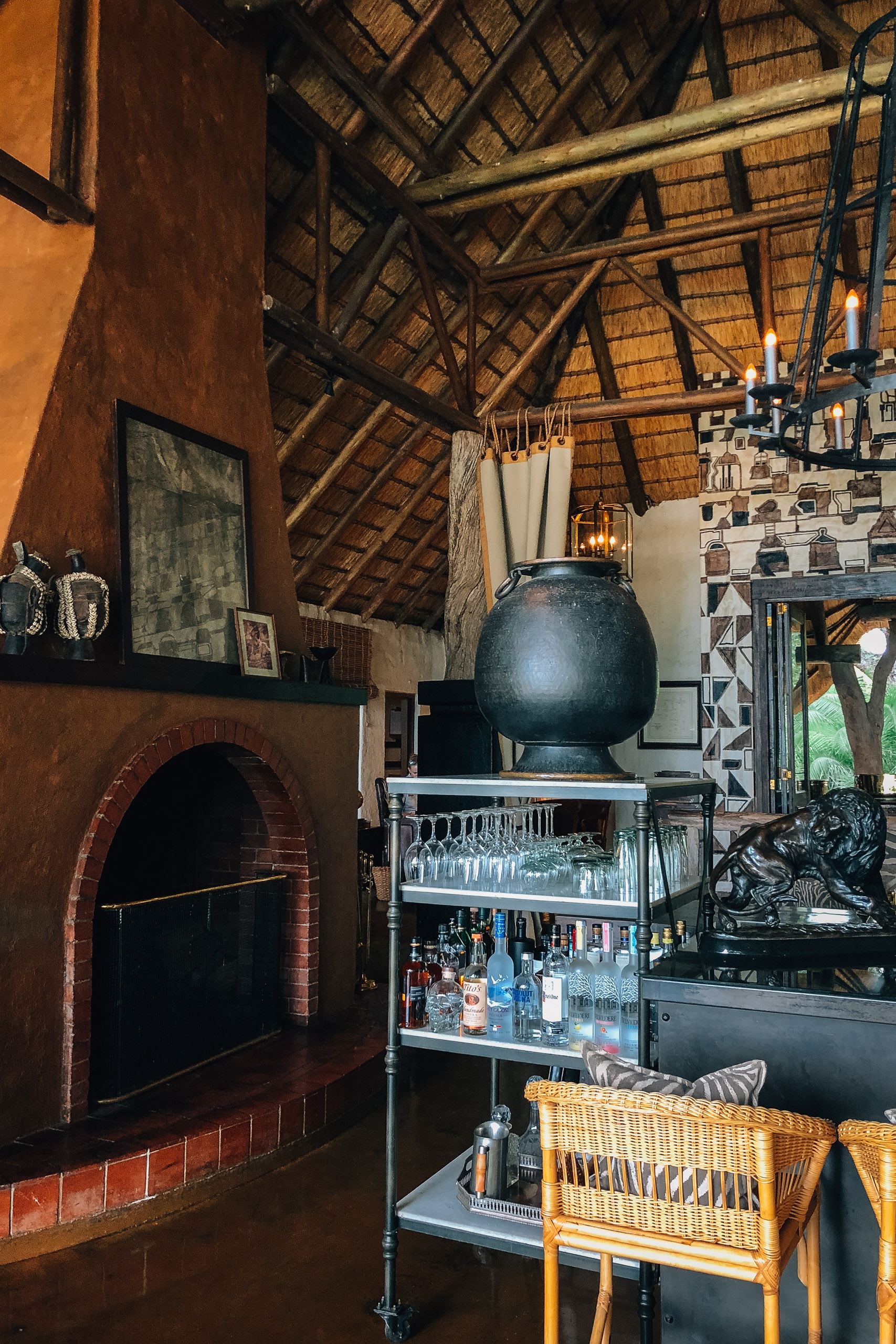 A rustic safari lodge interior features a red-brick fireplace, exposed wooden beam ceiling, and shelving displaying spirits and glassware beneath a large black decorative sphere