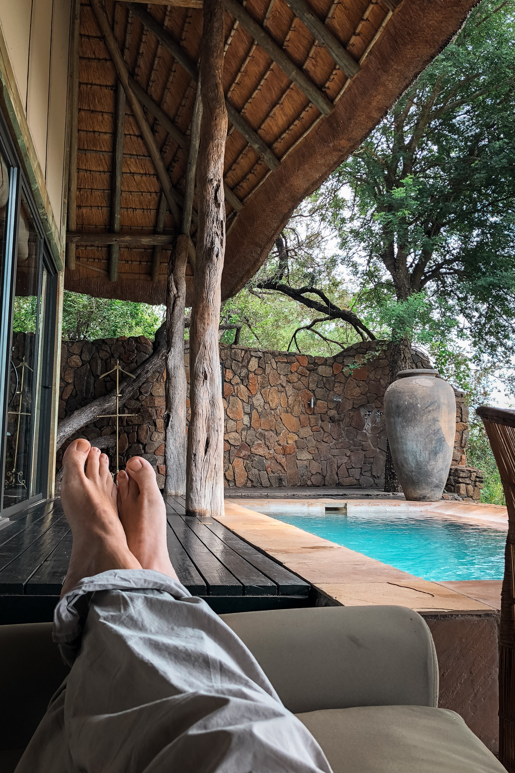 A person's bare feet rest on a wooden deck while relaxing under a thatched roof, overlooking a turquoise swimming pool and stone-walled lodge surrounded by lush green trees