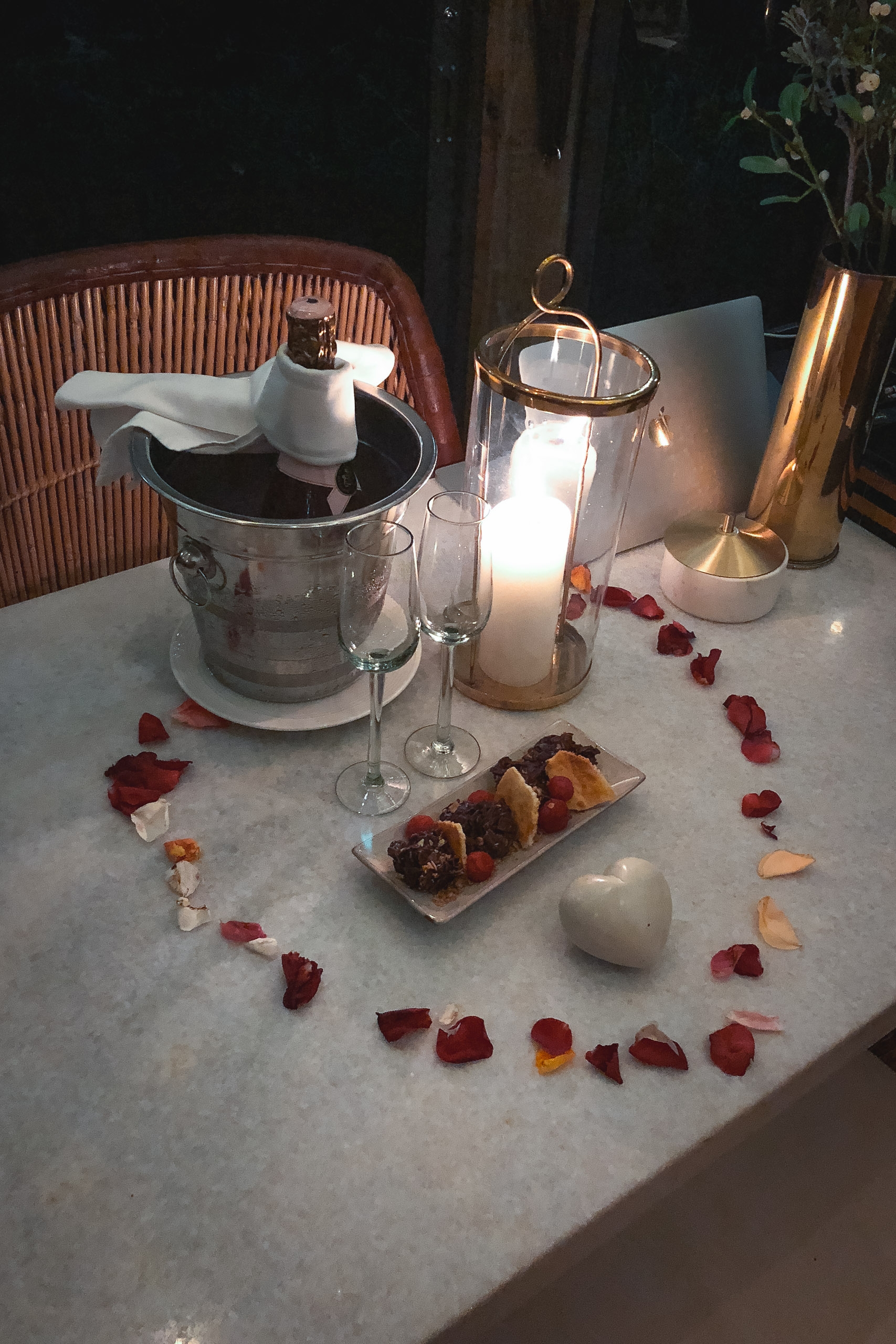 A marble tabletop arranged with champagne flutes, a white candle, brass vessels, pastries on a plate, and scattered rose petals forming a heart shape, bathed in warm natural light