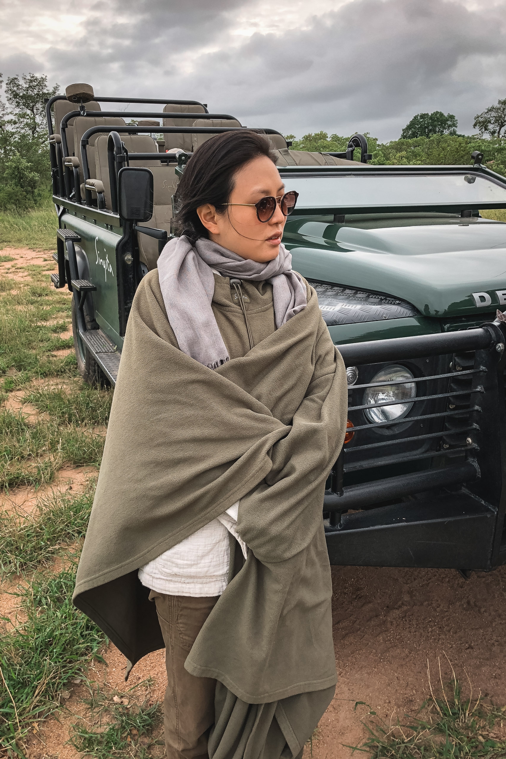 Woman in sunglasses and neutral layers stands wrapped in a tan blanket beside a green safari vehicle on a dusty African road under overcast skies