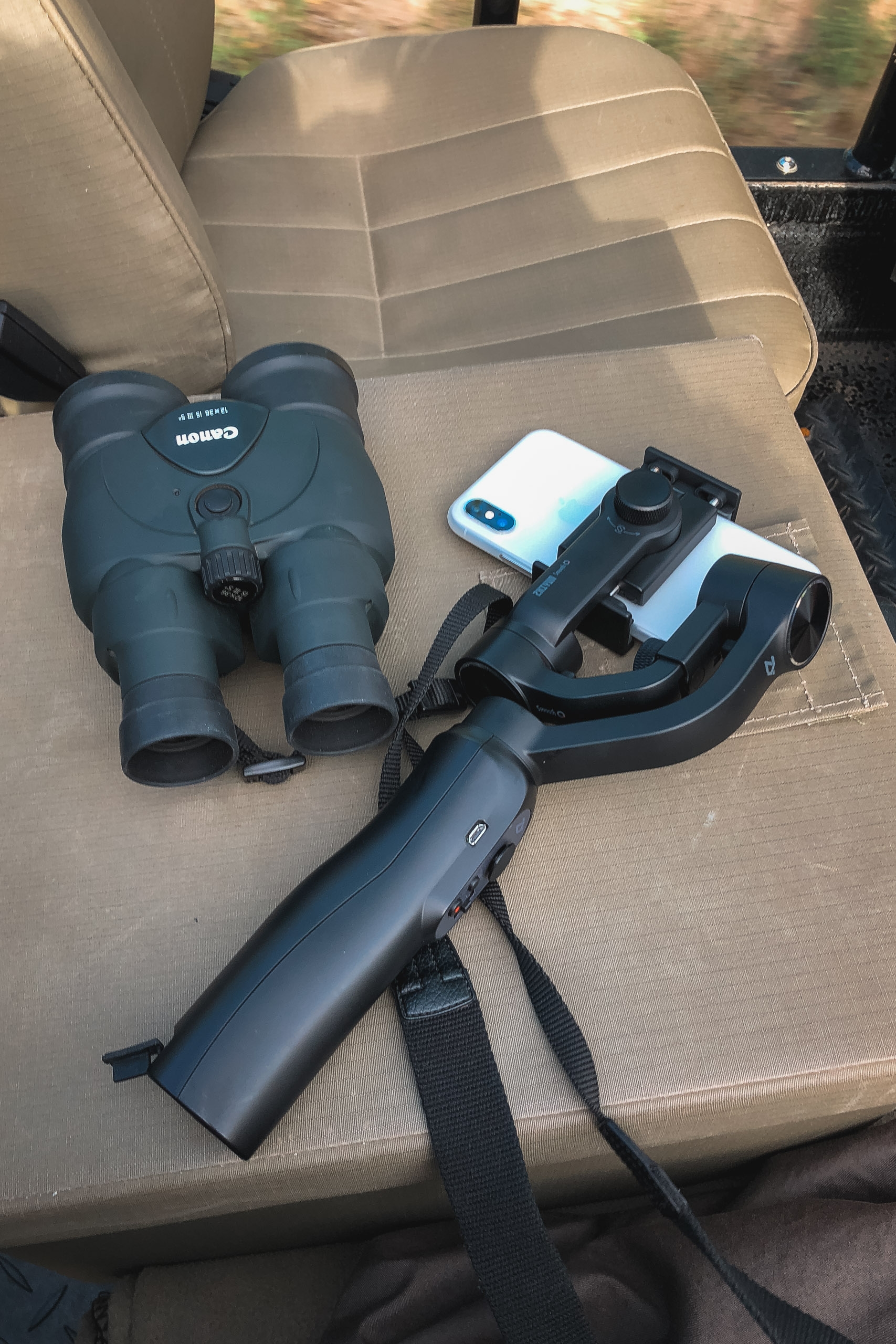 Blue binoculars and a smartphone gimbal stabilizer rest on a beige car seat during a safari outing in South Africa