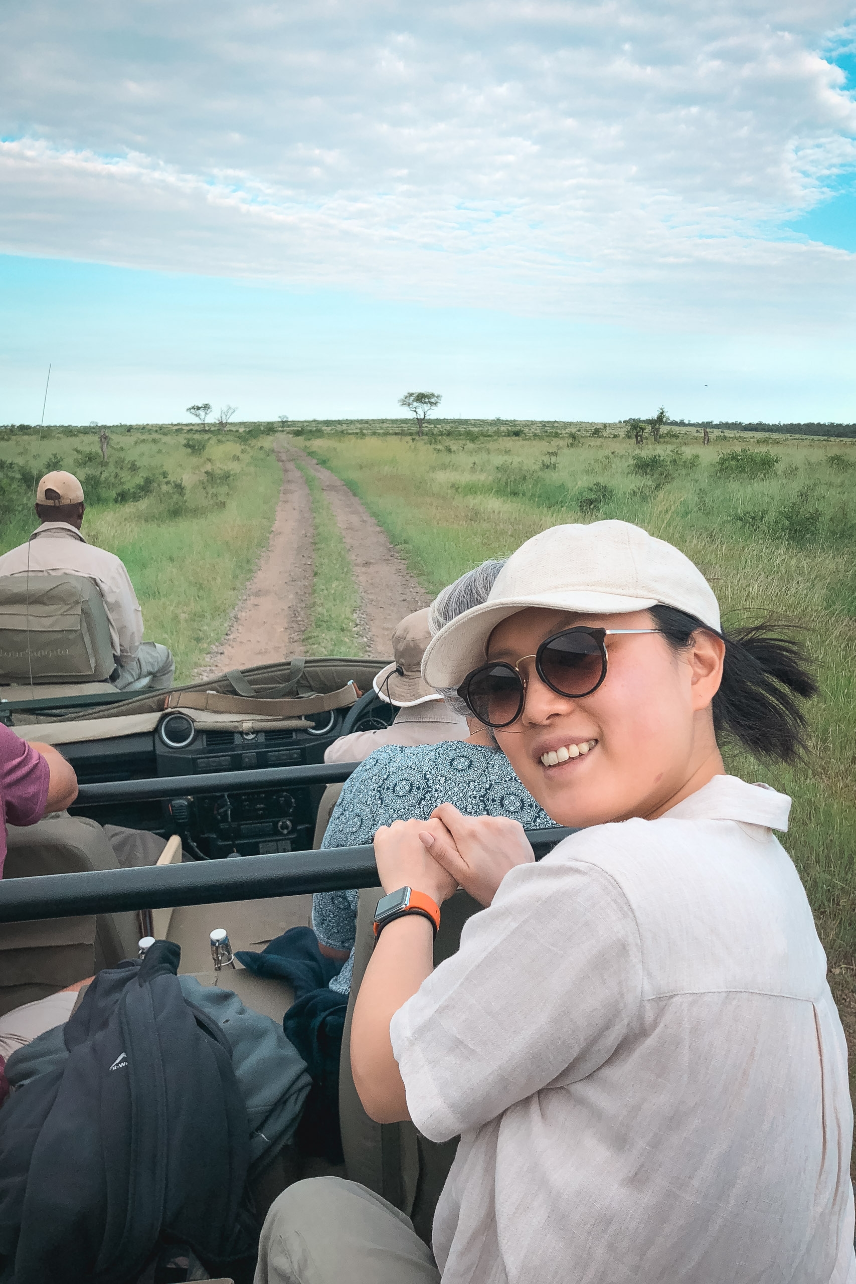 A smiling woman wearing a white cap and sunglasses sits in an open safari vehicle on a dirt road stretching across vast green grassland under a partly cloudy sky
