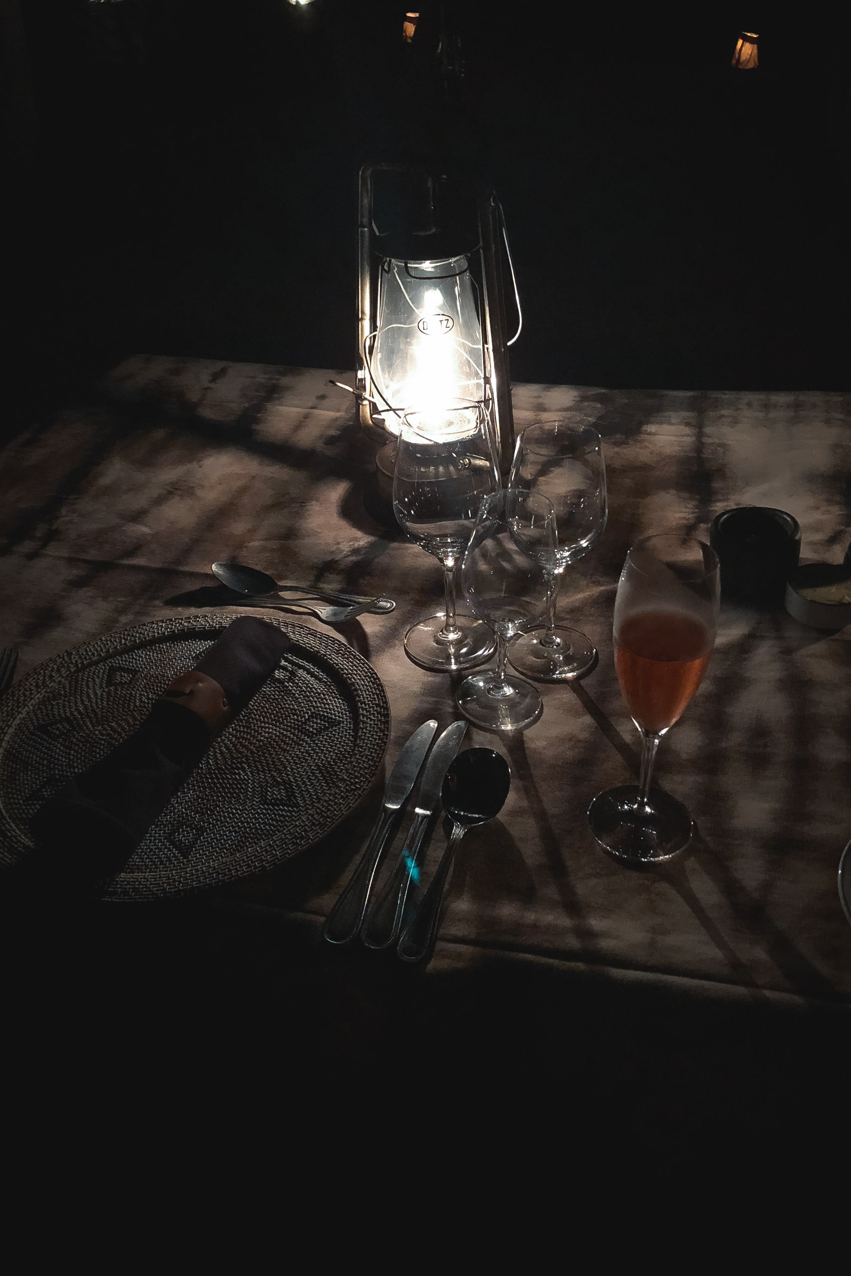 A formal outdoor dinner table set with glowing lantern, empty wine glasses, folded napkin, and cutlery, bathed in dramatic shadows at night