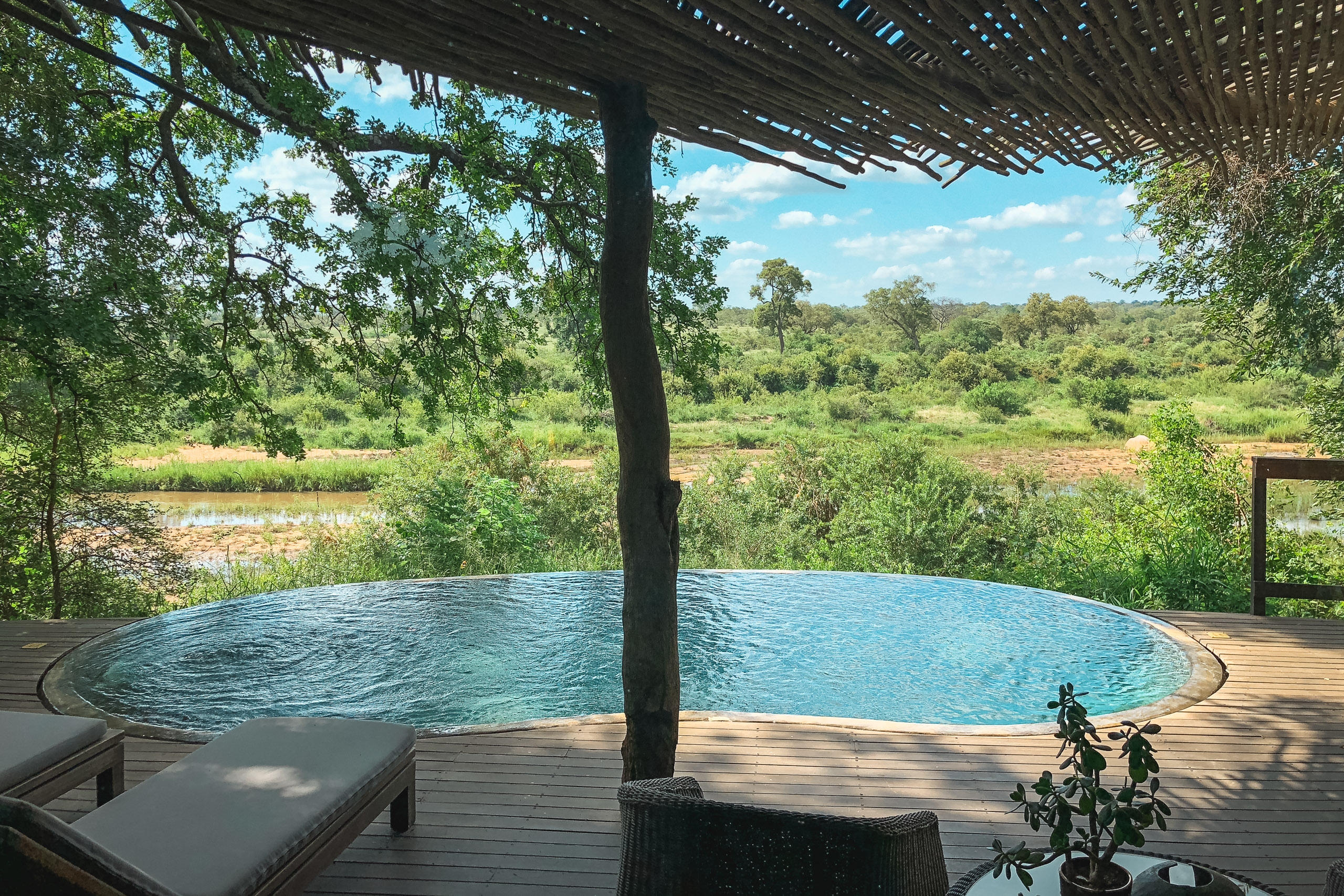 A circular infinity pool on a wooden deck overlooks a lush riverine landscape, with a natural river visible beyond and dense green vegetation stretching toward a blue sky
