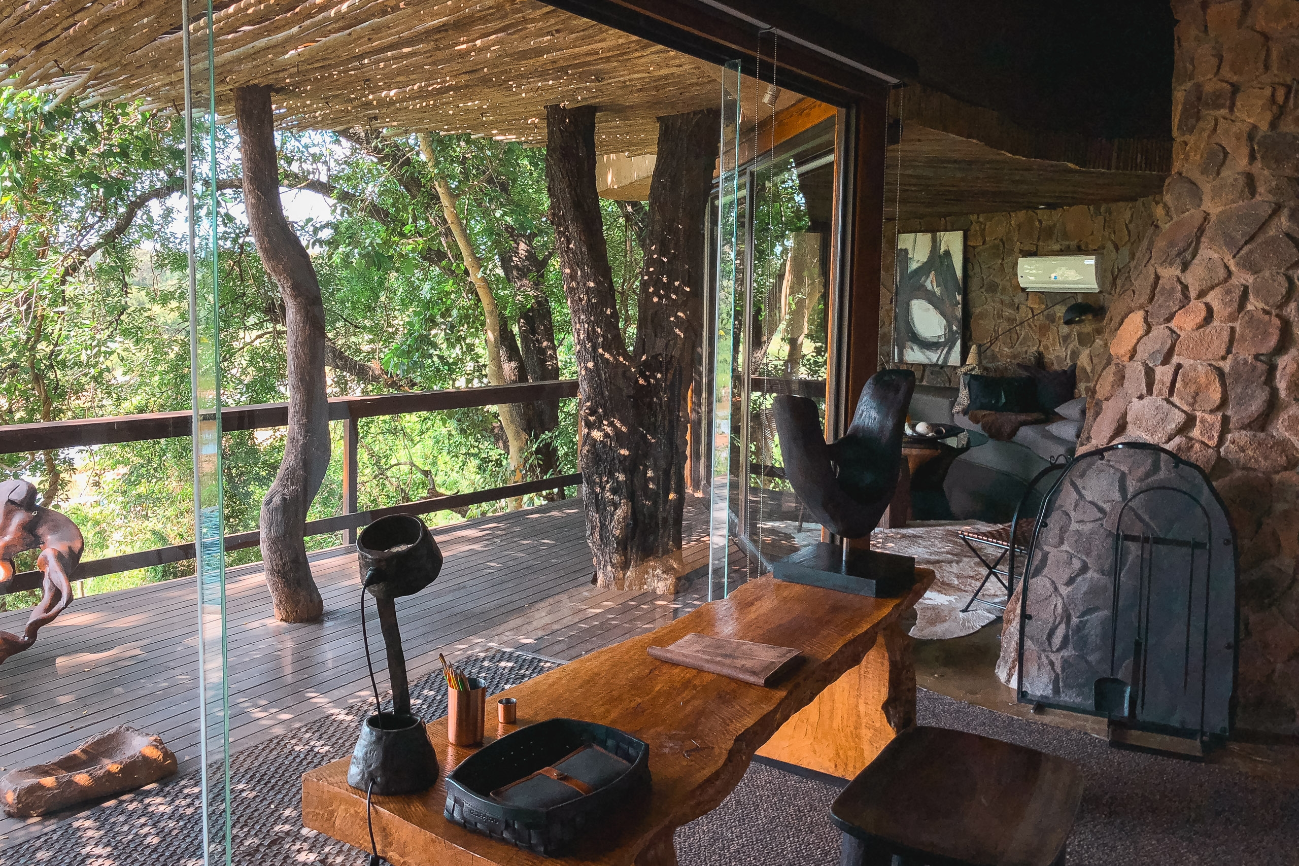 A rustic safari lodge interior opens through glass doors onto a wooden deck where gnarled tree trunks frame views of dense green foliage, with stone walls and furnishings visible inside the naturally lit space