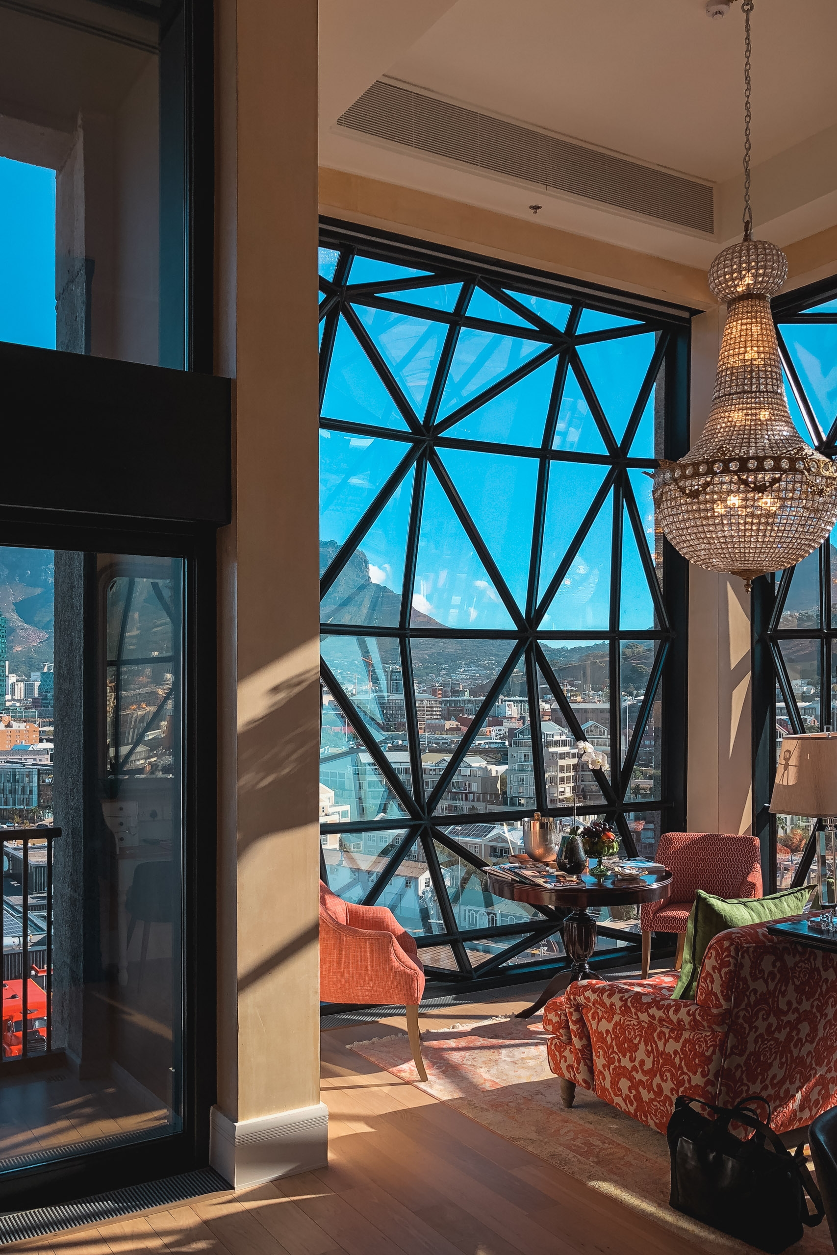 High-rise hotel suite with geometric black-framed windows overlooking Cape Town's cityscape, featuring coral upholstered chairs, a crystal chandelier, and warm sunlit interiors