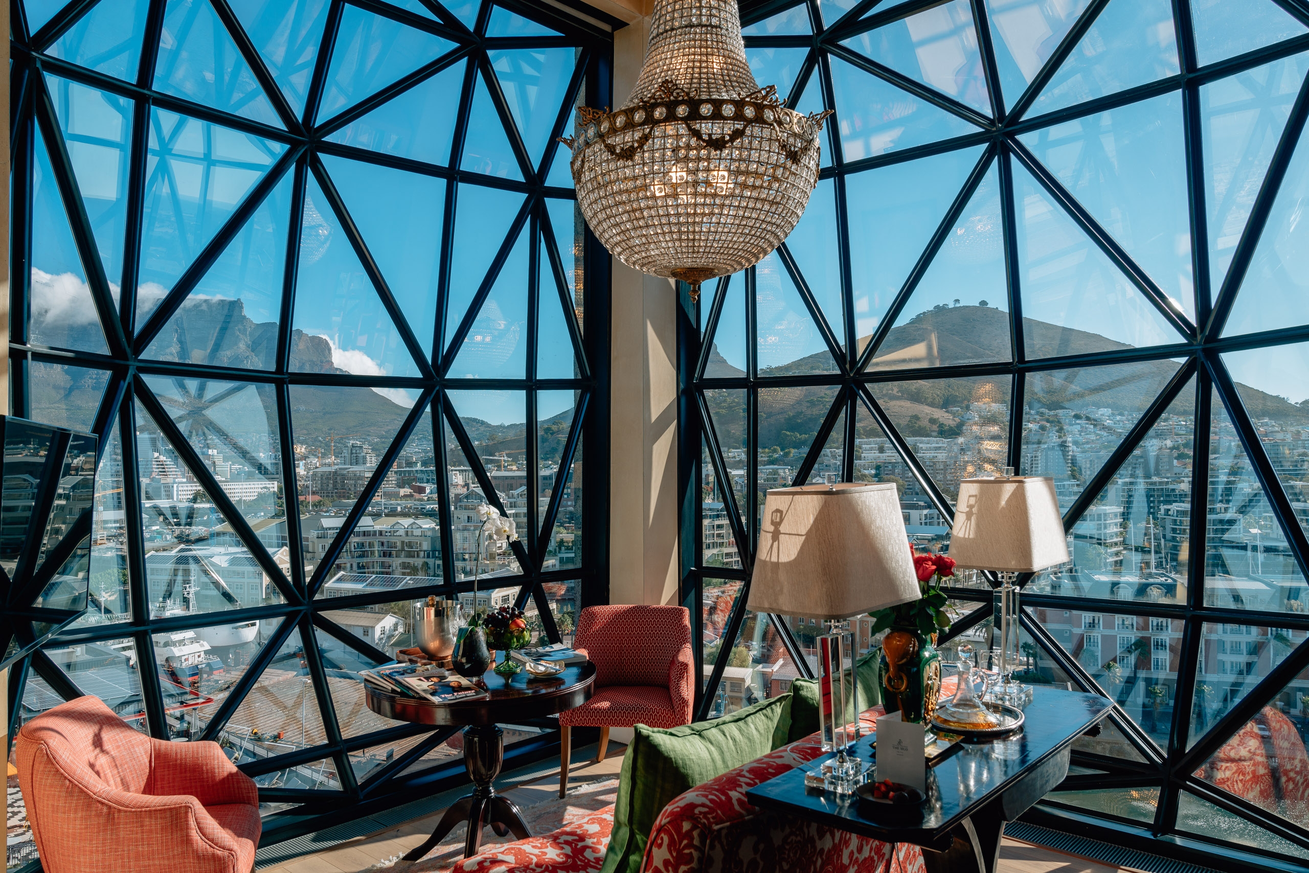 A high-rise hotel lounge features geometric gridded windows framing panoramic city and mountain views, with a beaded chandelier, colorful furnishings, and contemporary décor bathed in natural daylight