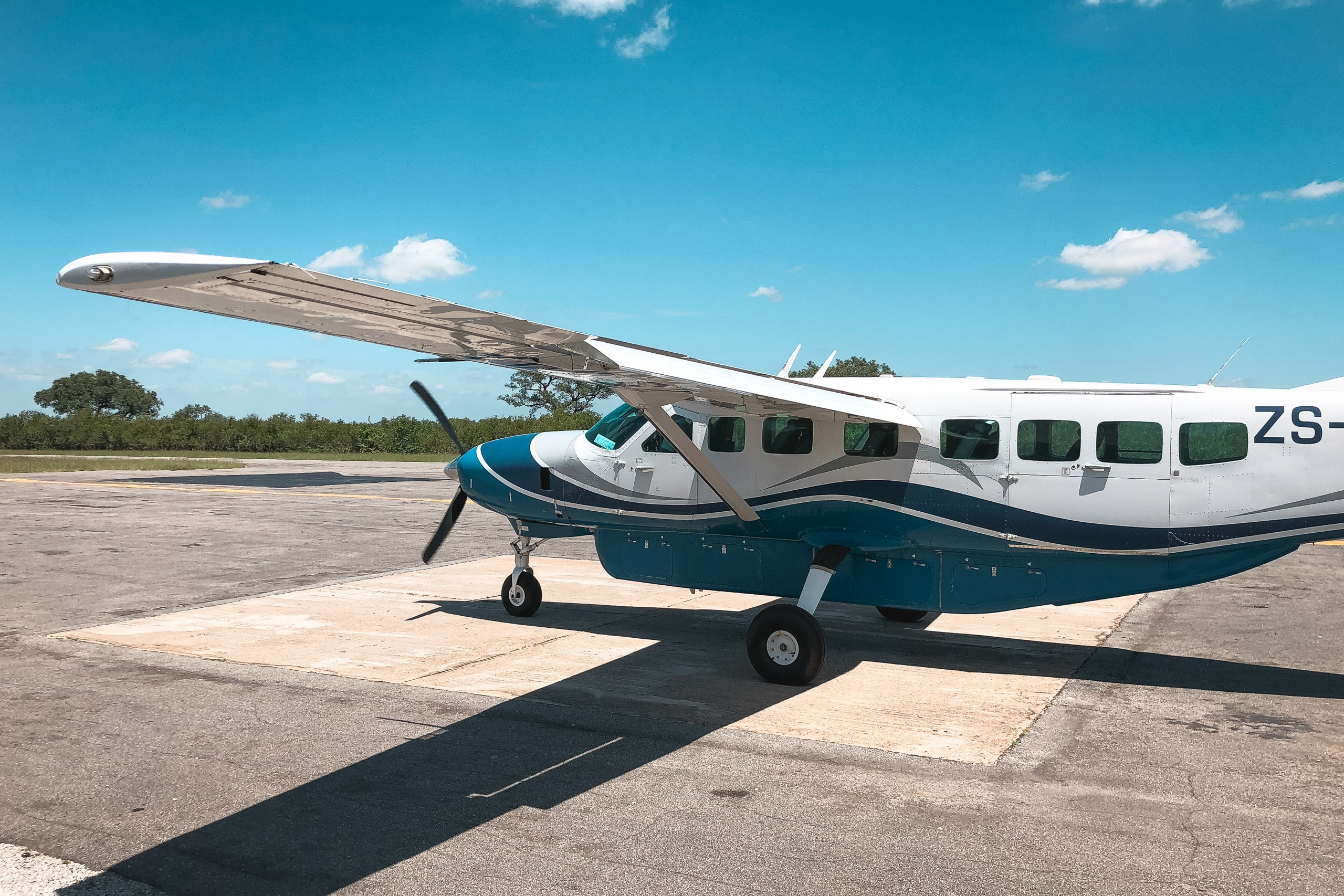 A teal and white Cessna passenger aircraft sits parked on a concrete runway under clear blue sky with sparse clouds and distant tree line visible
