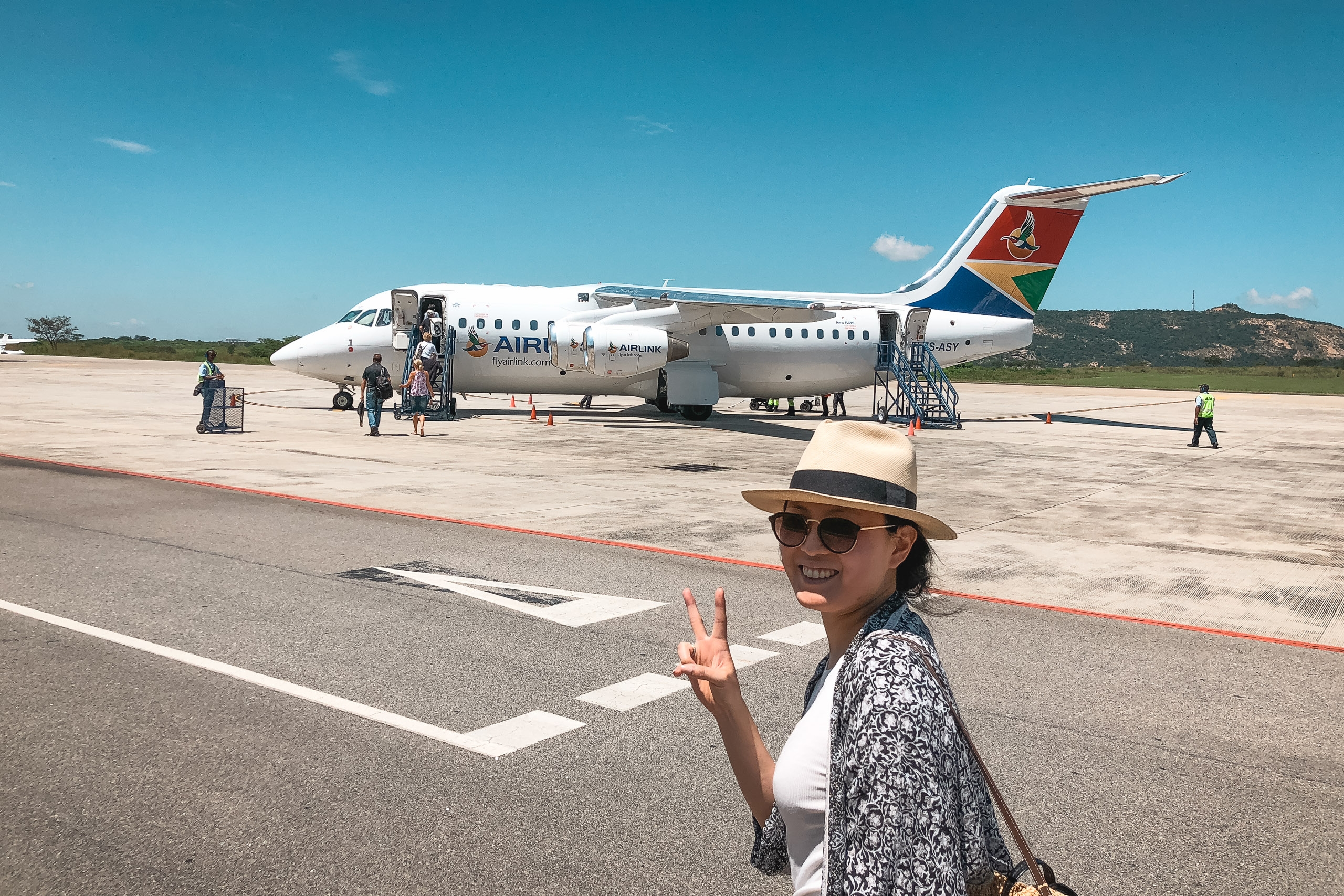 A woman in a straw hat and sunglasses makes a peace sign on a tarmac as an Air Namibia aircraft prepares for departure under clear blue sky