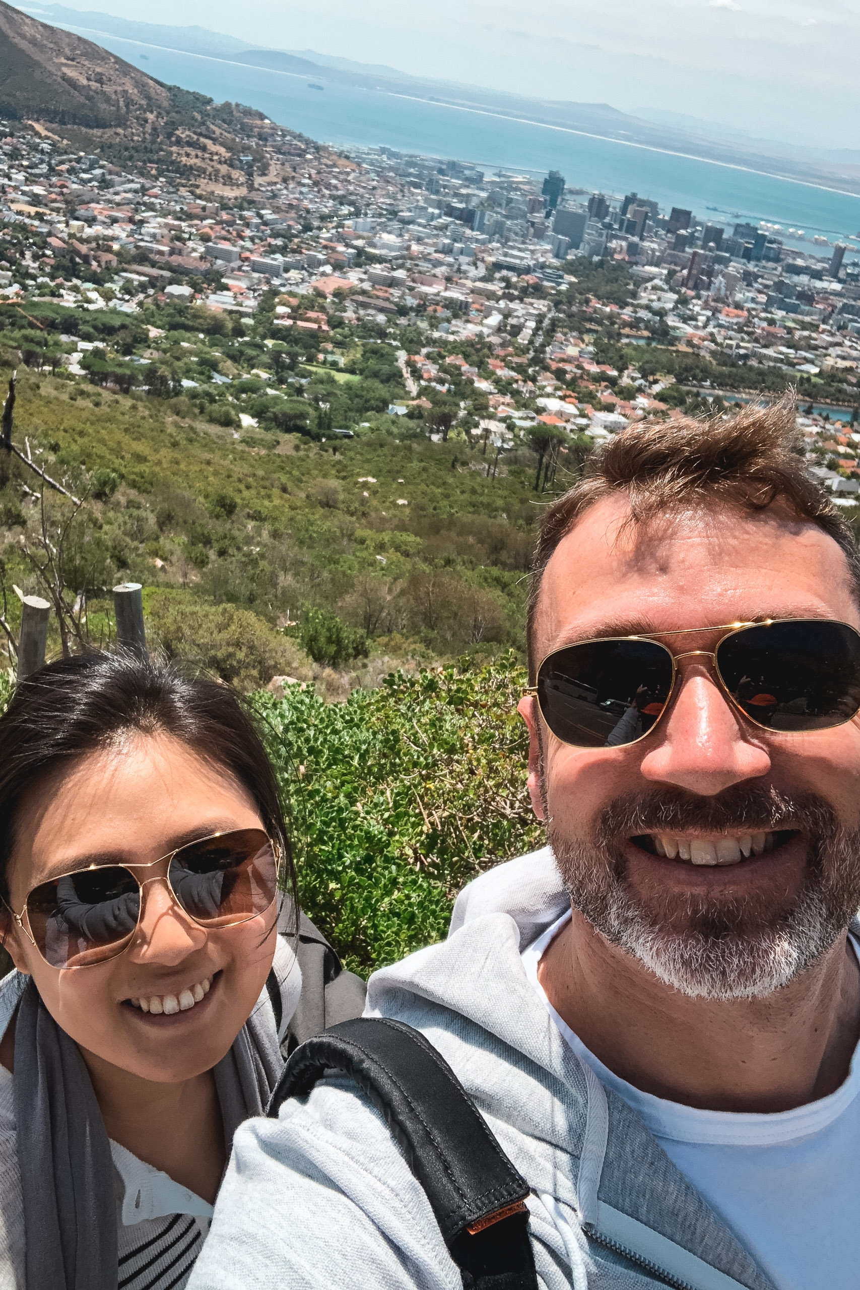 Two smiling visitors in sunglasses pose on a mountain overlook with Cape Town's sprawling cityscape, turquoise coastline, and distant peaks visible beneath bright afternoon sunlight