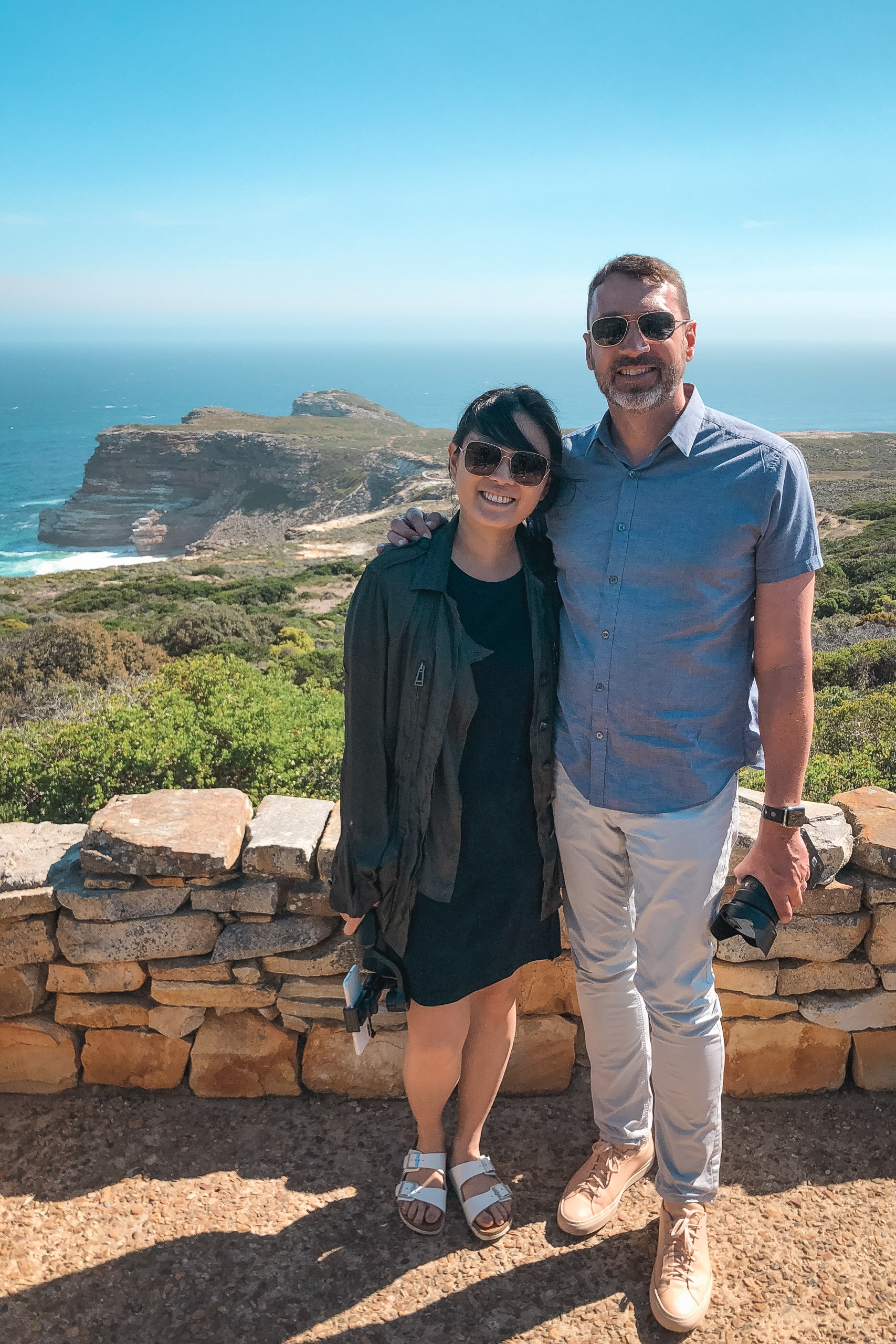 A couple stands together at a stone overlook wearing sunglasses, with a dramatic coastal headland and turquoise ocean visible behind them under clear blue skies
