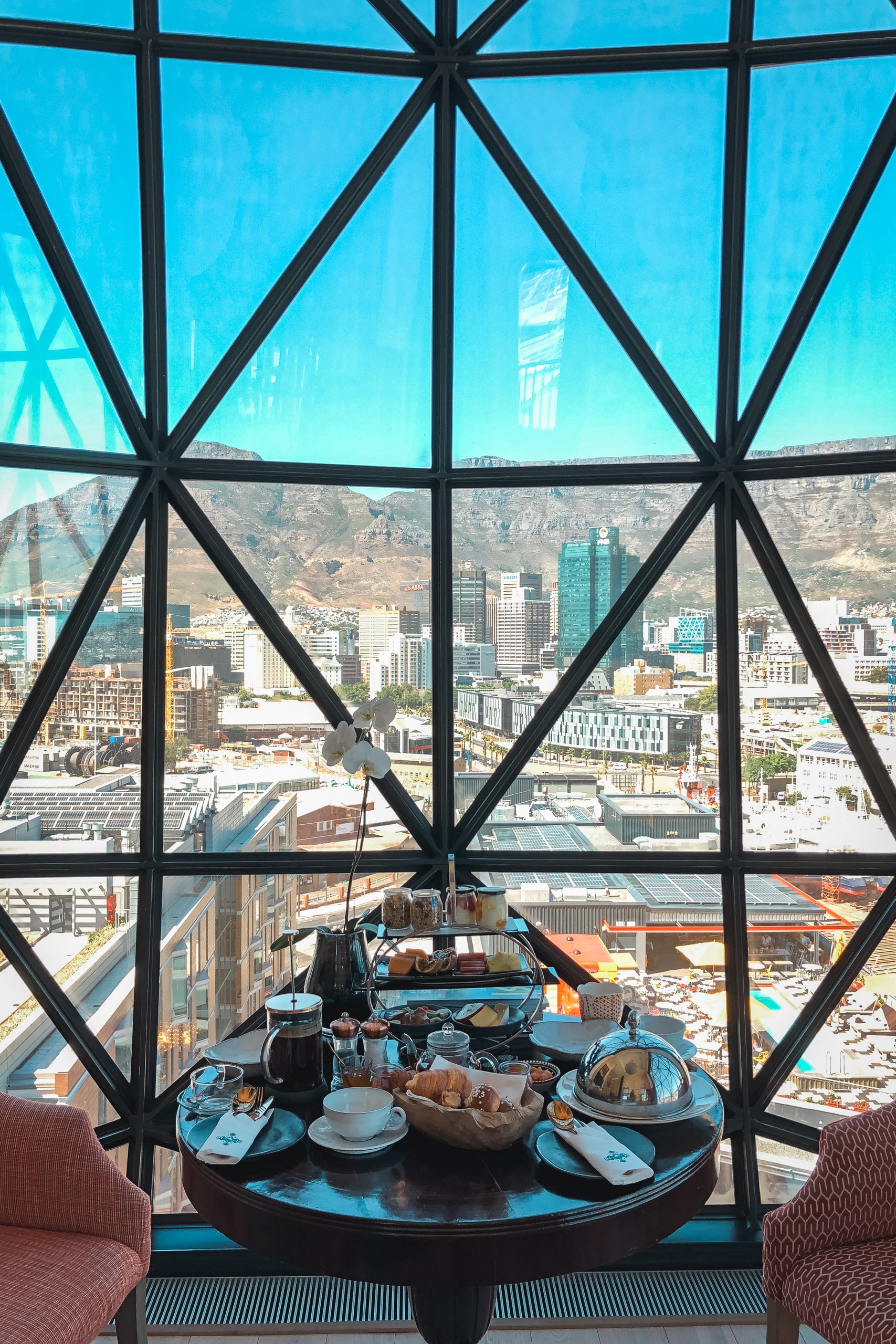 A high-altitude dining table with place settings overlooks Cape Town's cityscape and Table Mountain through geometric black-framed windows on a clear day
