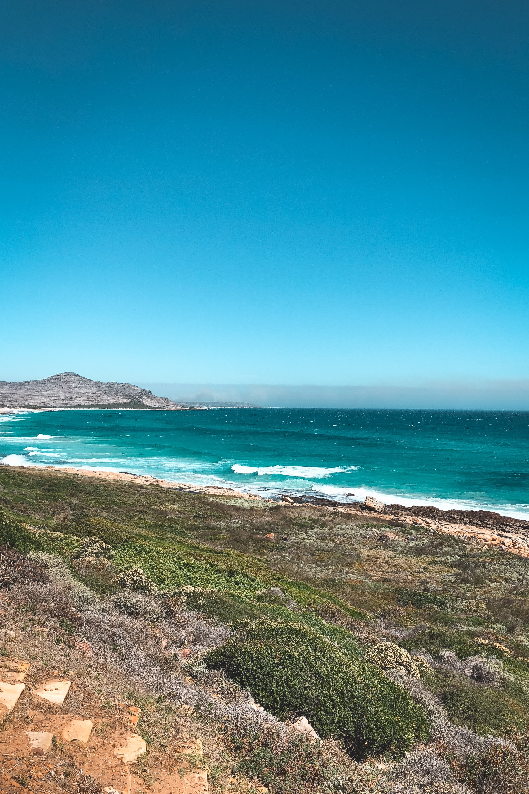 Coastal landscape with turquoise waves rolling onto a sandy beach, rocky headland covered in sparse vegetation, and a distant mountain under clear blue sky