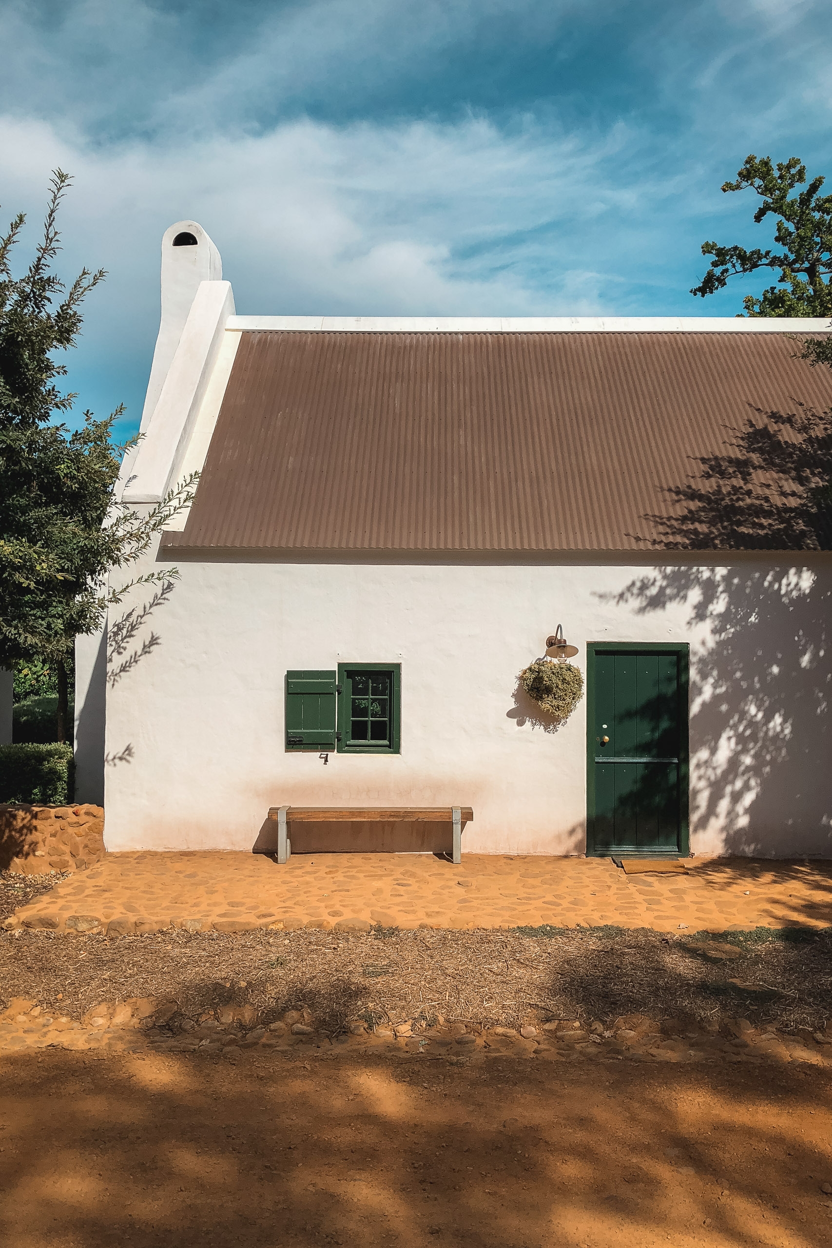 White-walled cottage with green shutters and door sits on reddish earth beneath dramatic cloudy sky, casting sharp tree shadows across the sandy courtyard