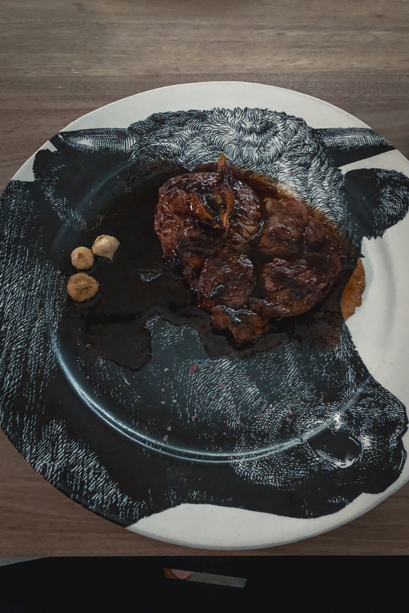 A decorative white plate with an etched black bull's head design holds a seared steak and scattered mushrooms, set against a wooden surface