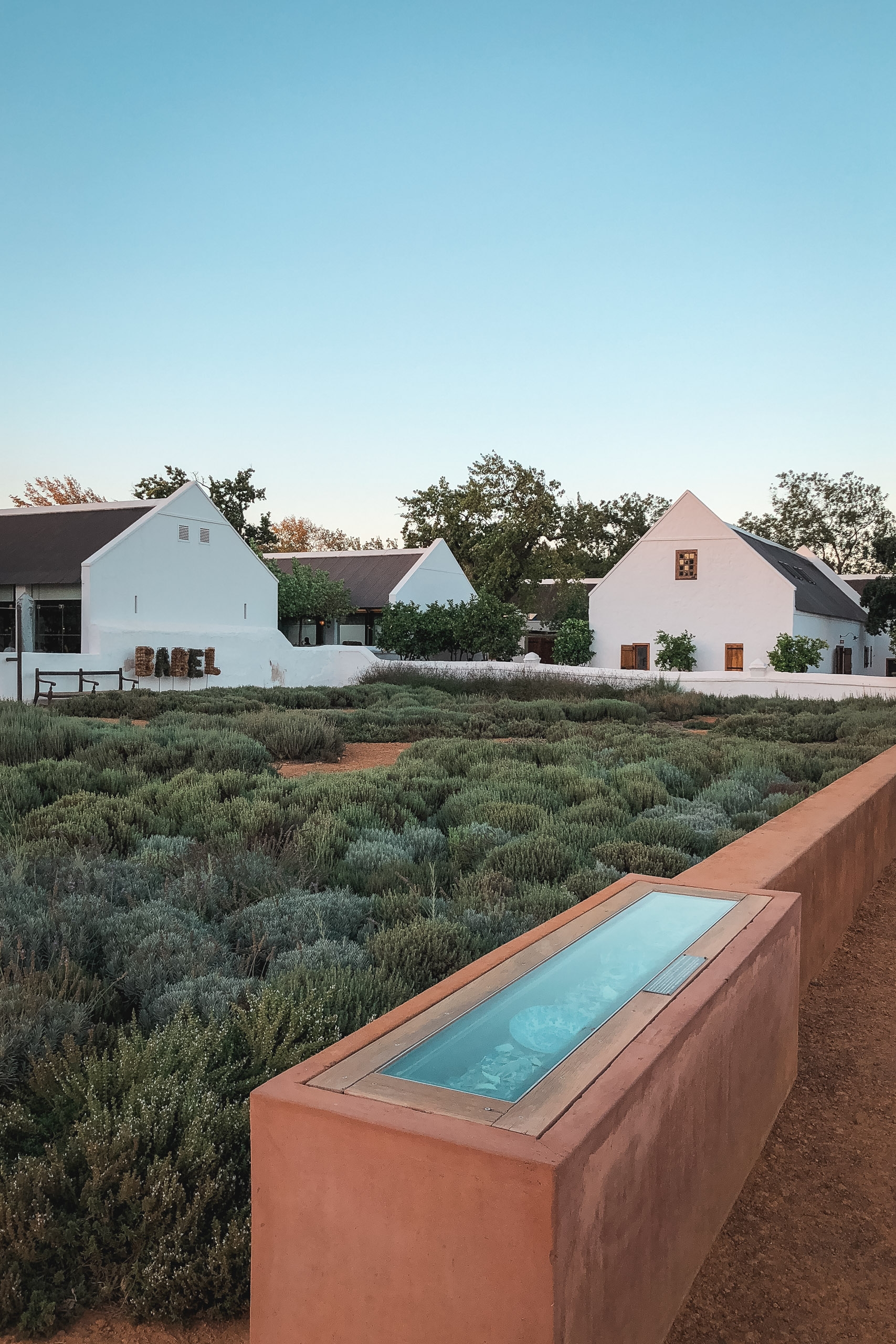 A terracotta water feature sits in the foreground of a sparse, drought-resistant garden overlooking white-walled farm buildings with gabled roofs under clear blue sky