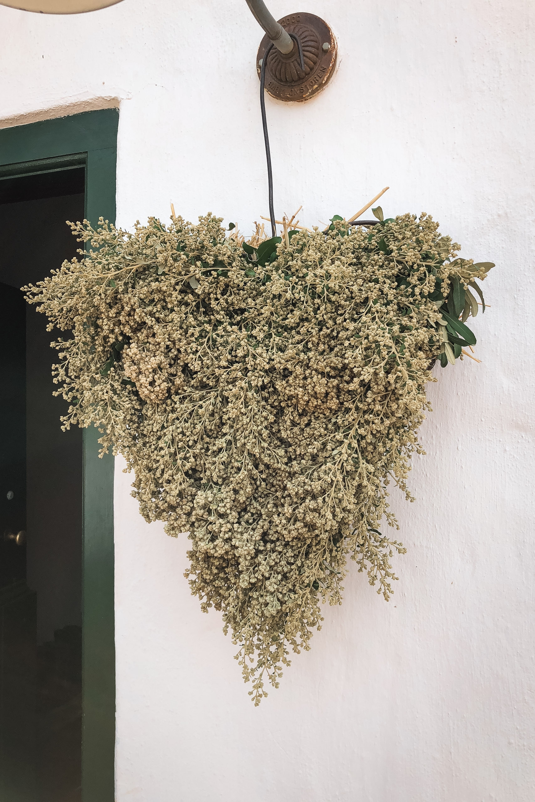 A lush hanging basket of white baby's breath cascades from a decorative brass wall mount beside a green-framed doorway on a whitewashed exterior wall