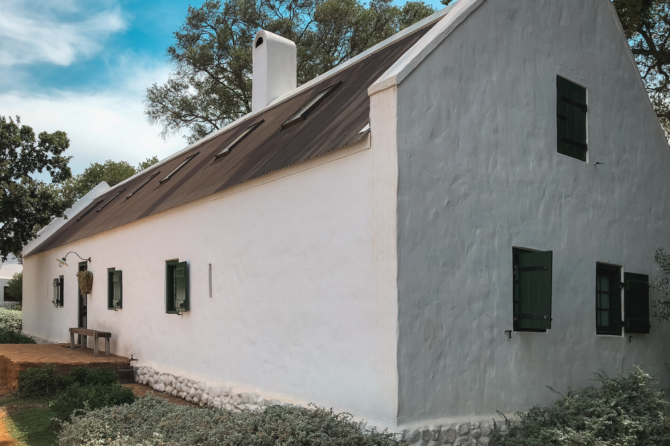 A whitewashed farmhouse with a dark thatched roof stands against blue sky, surrounded by green vegetation and mature trees under bright daylight conditions