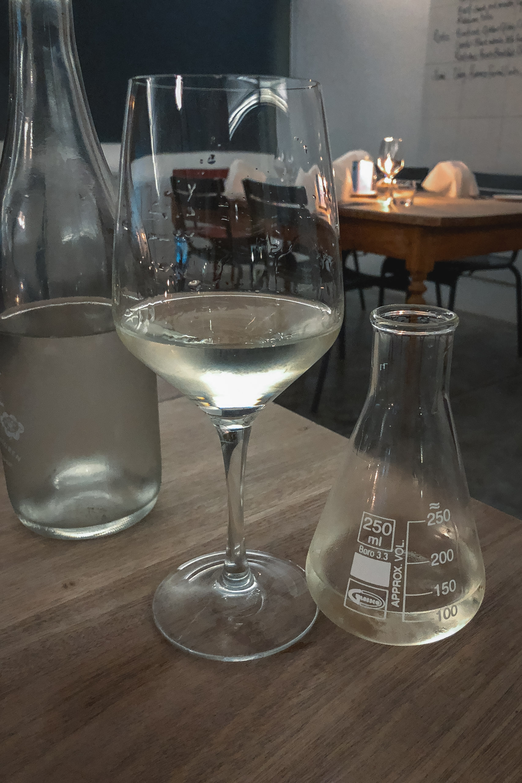 Wine glass filled with pale liquid sits beside laboratory glassware and a bottle on a wooden table, with blurred interior setting and warm lighting in background