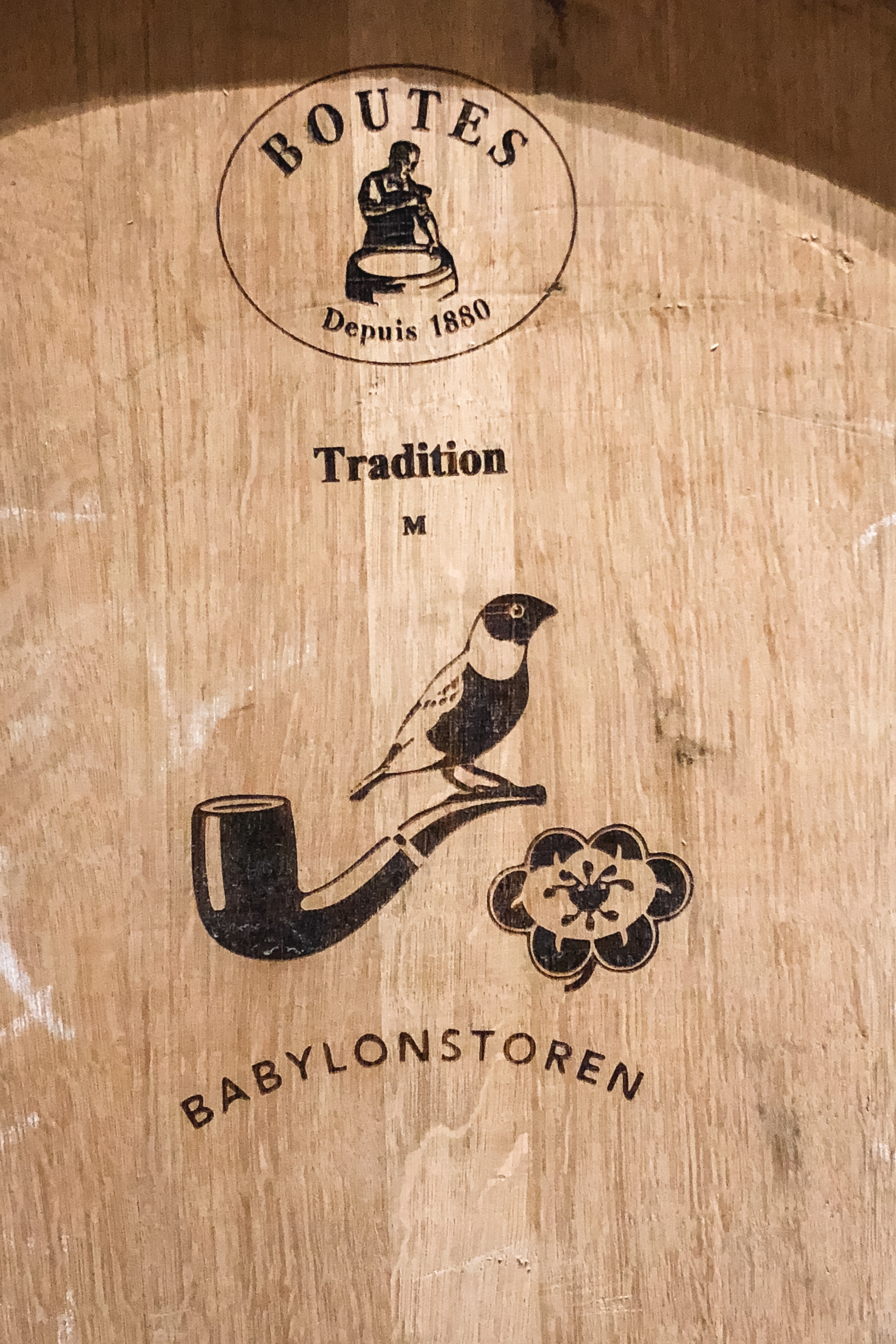 Wooden barrel lid featuring the "Boutes" logo from 1860, with "Tradition M" text and decorative bird and pipe illustrations above "Babylonstoren" branding