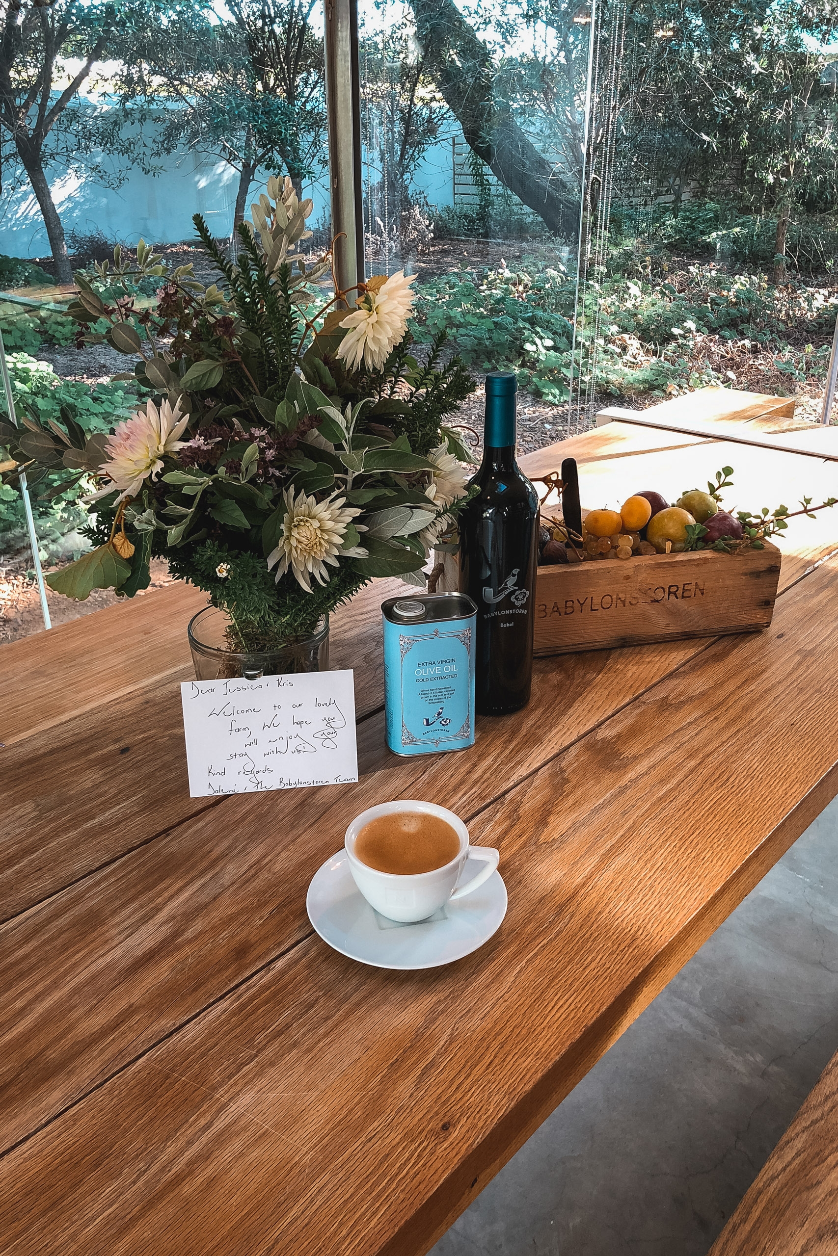 A wooden table displays a white espresso cup, fresh daisies in a vase, a wine bottle, citrus fruits in a wooden crate, and a handwritten note, overlooking a sunlit garden through large windows