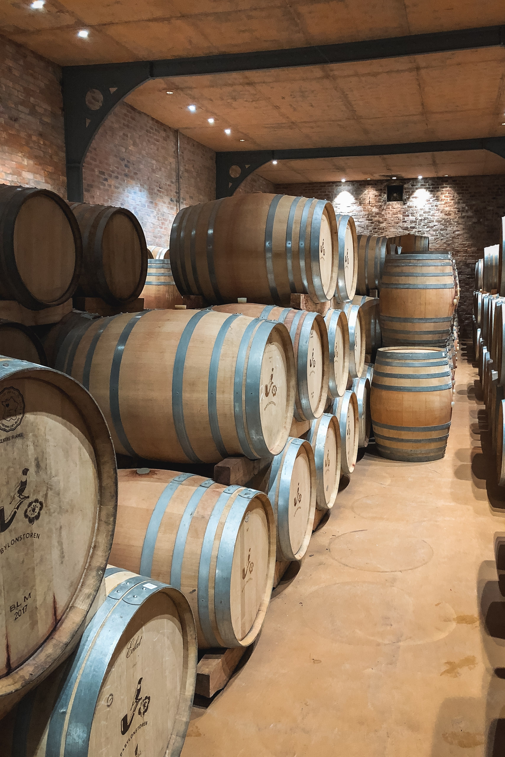 Wooden wine barrels with metal bands stacked in rows inside a brick winery cellar with industrial lighting and exposed beam ceiling