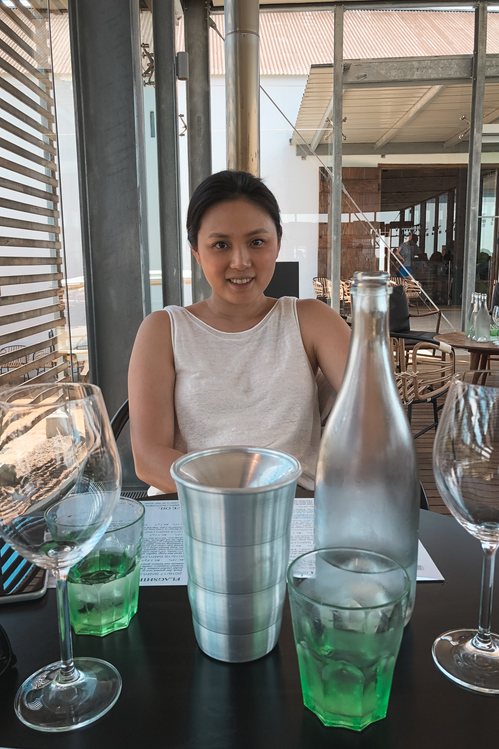 A smiling woman in a white sleeveless top sits at a table with green cocktails and metallic drinkware, framed by modern glass windows and bright natural light from an open-air venue