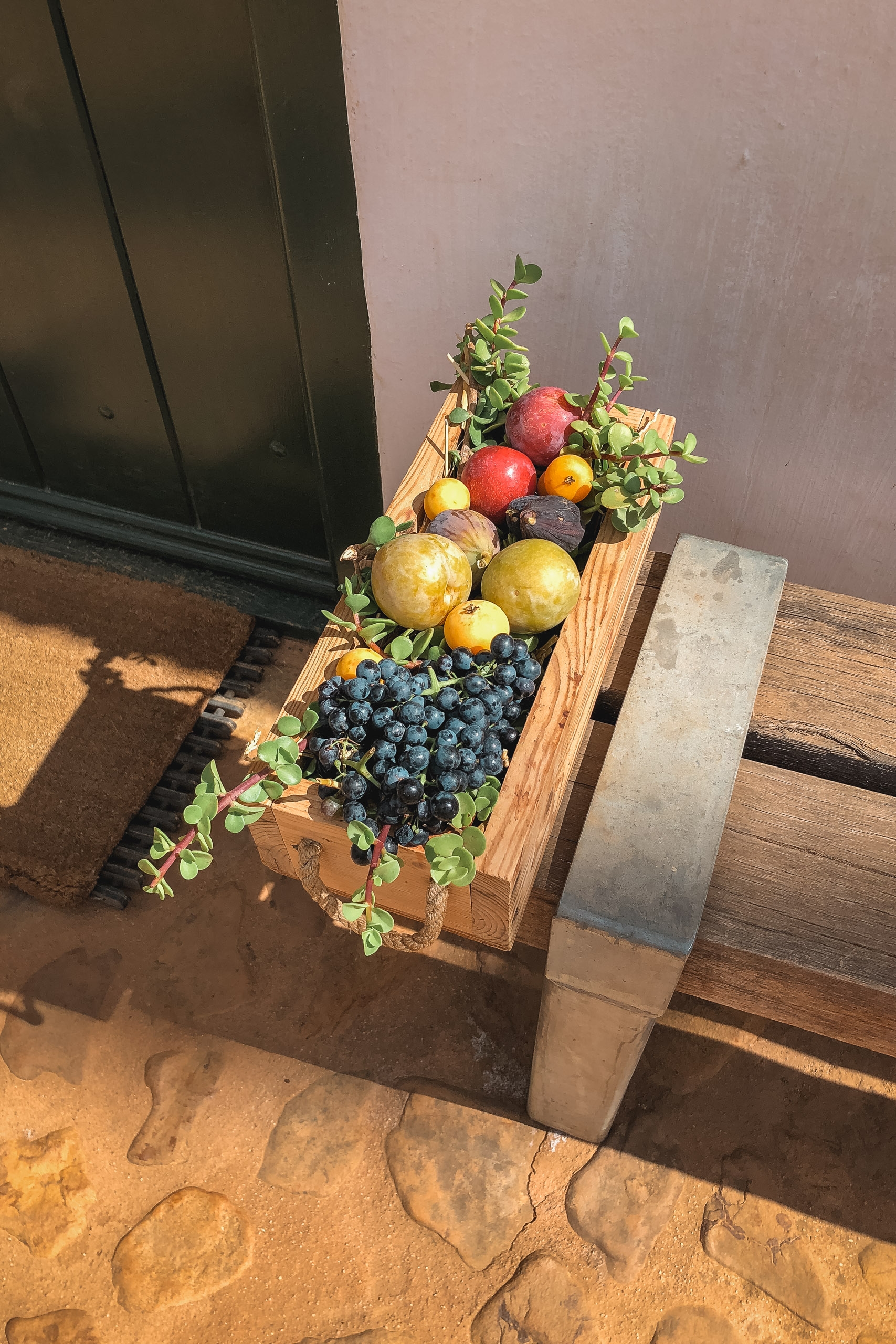 A wooden crate overflowing with fresh produce—red apples, yellow pears, dark blueberries, and green foliage—sits on a wooden deck beside a modern building in warm sunlight