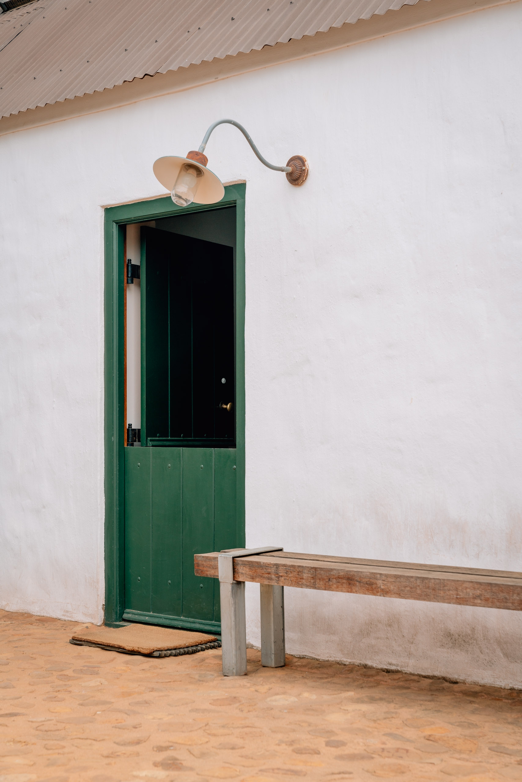 A weathered white wall features a green-framed door with a curved copper lamp mounted above it, beside a wooden bench on sandy ground