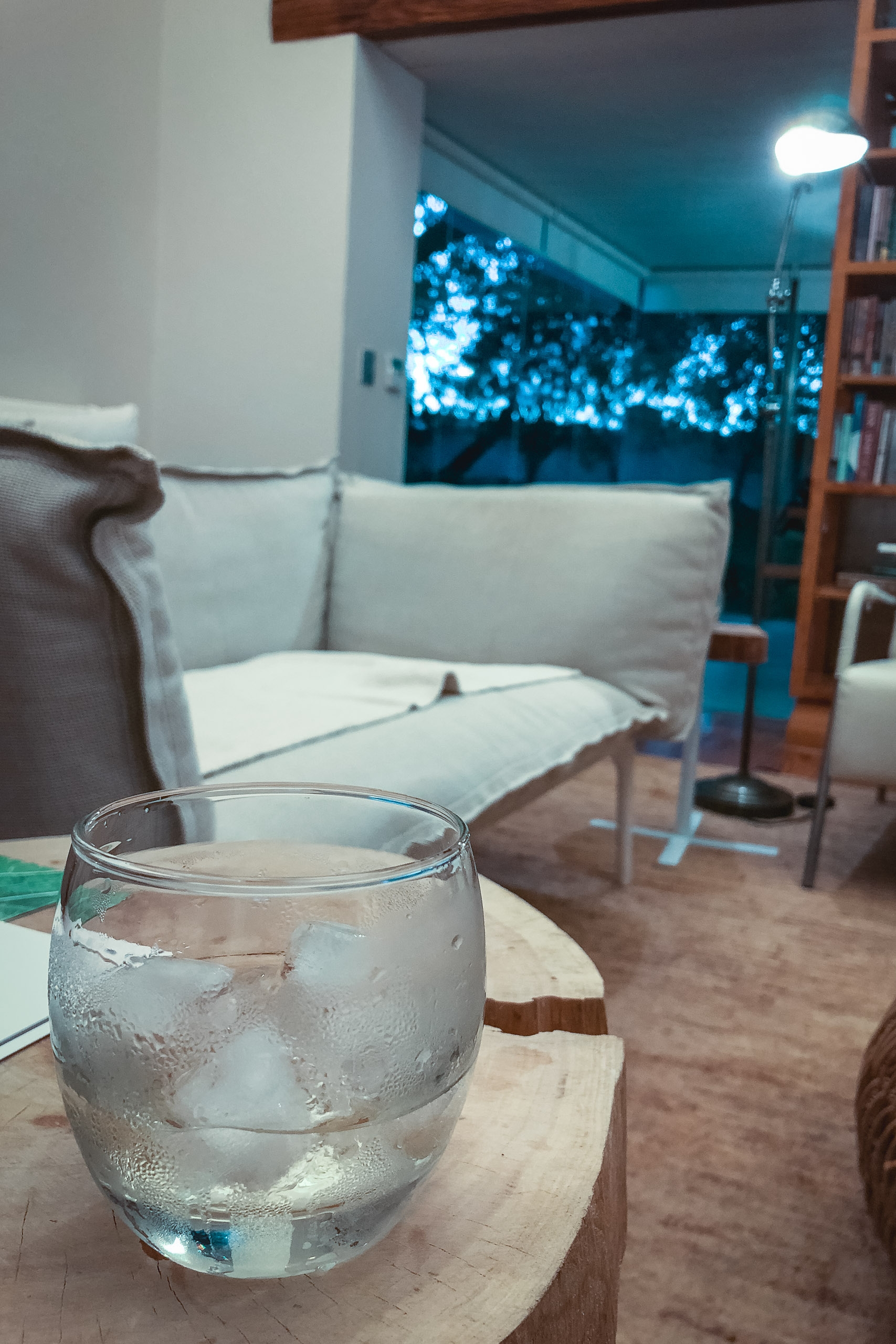 A glass of ice water sits on a wooden table in a modern living room with a cream sofa, warm overhead lighting, and blue bokeh lights visible through the window at dusk