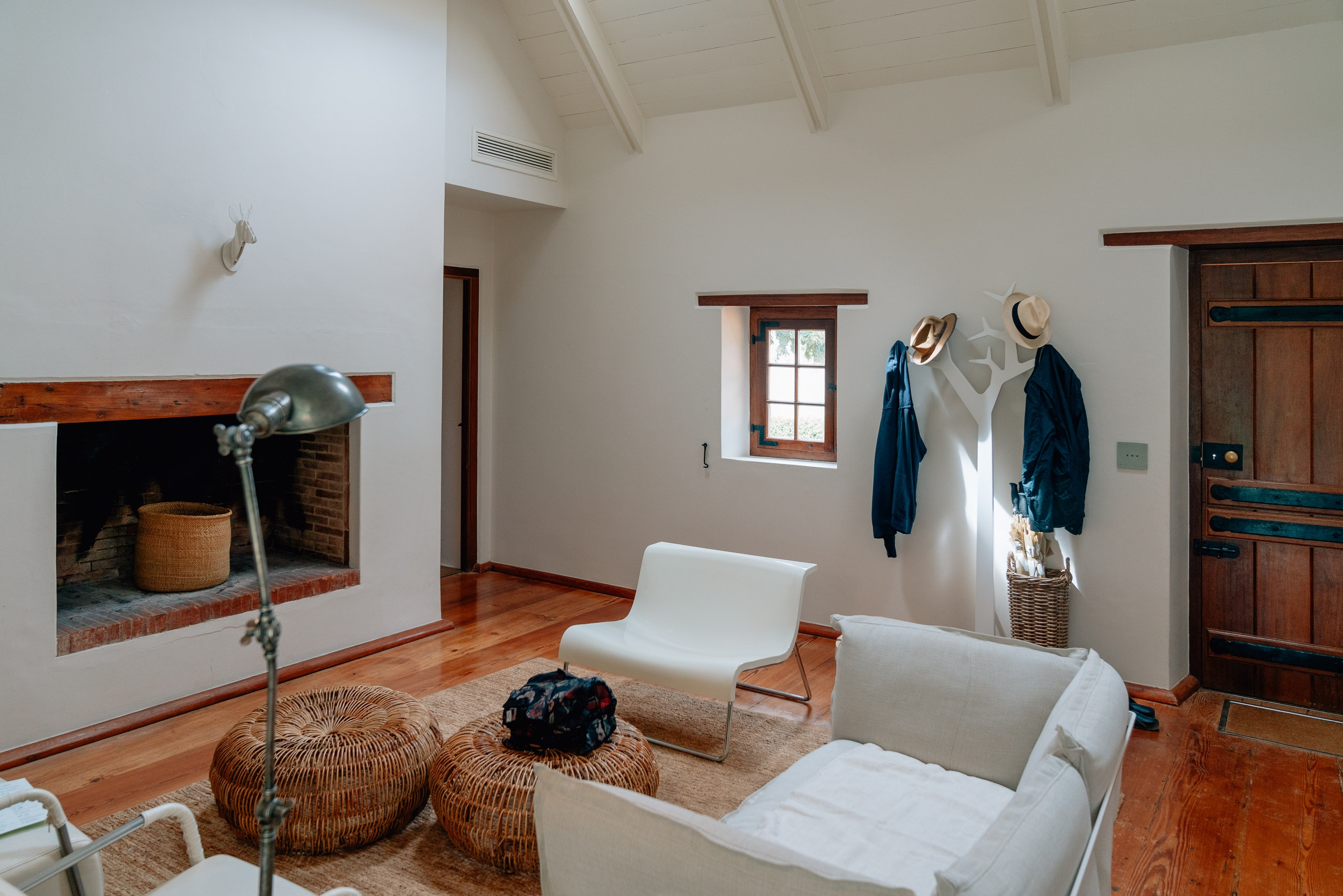 A minimalist bedroom interior features cream-colored walls, wooden beams, and polished wood floors, with a white bed, wicker accessories, and clothing hung on a white wall rack near a small window