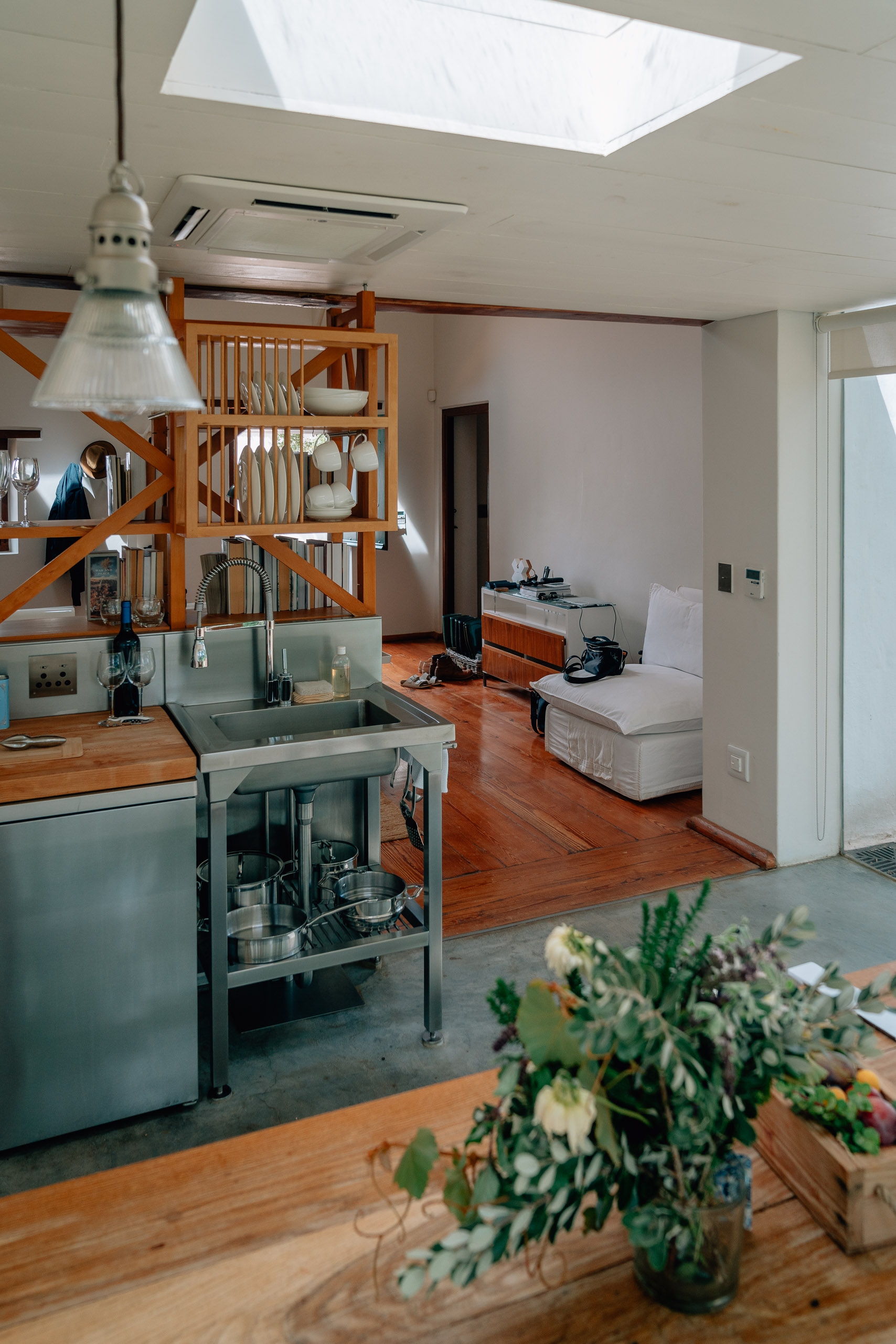 A modern open-plan kitchen and living space features warm wooden flooring, a green island sink with stainless steel appliances, wooden dish racks, and a skylight flooding the room with natural light