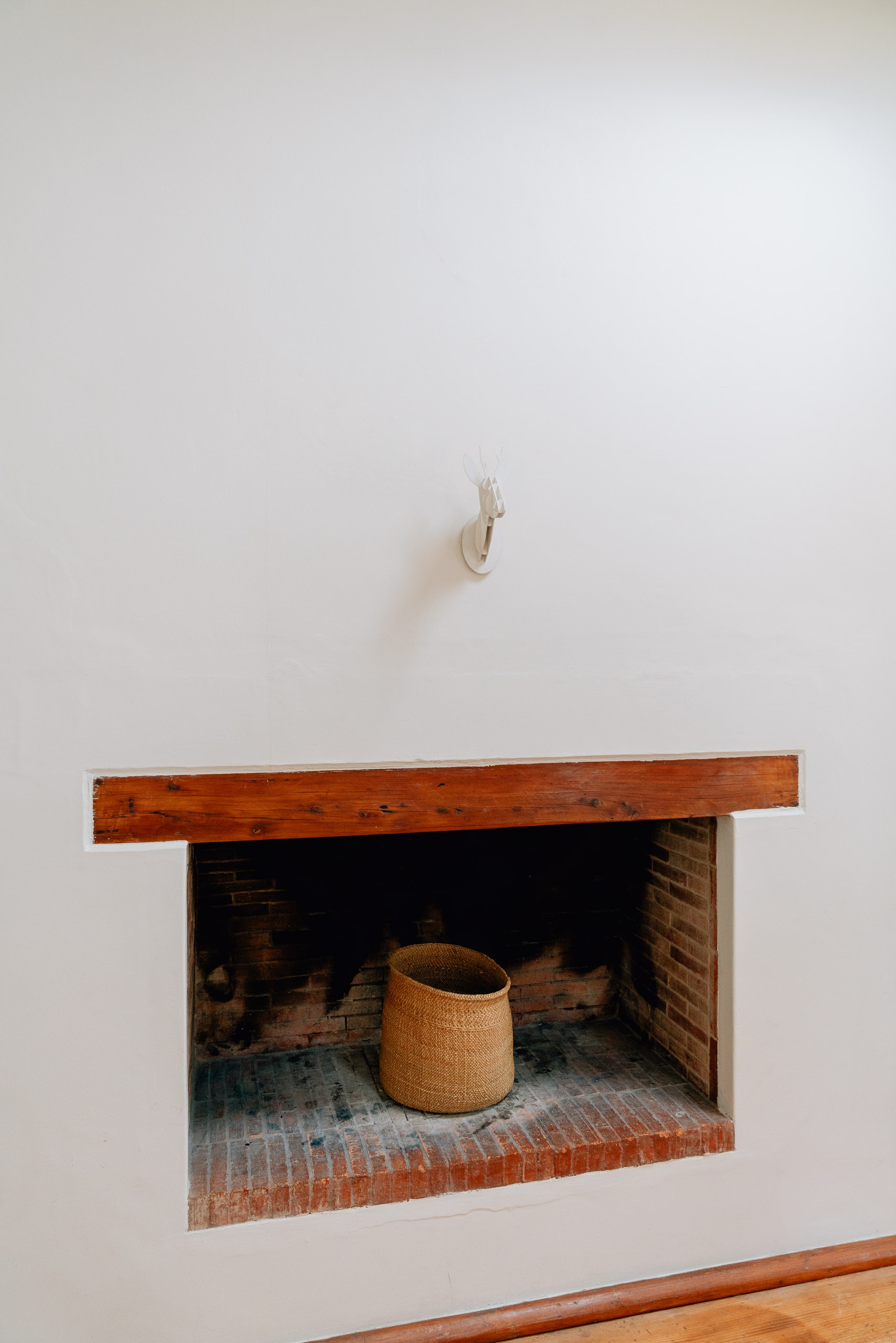 A rustic brick fireplace set into a white wall holds a woven basket, with a wooden mantel above and a hook mounted on the pale wall